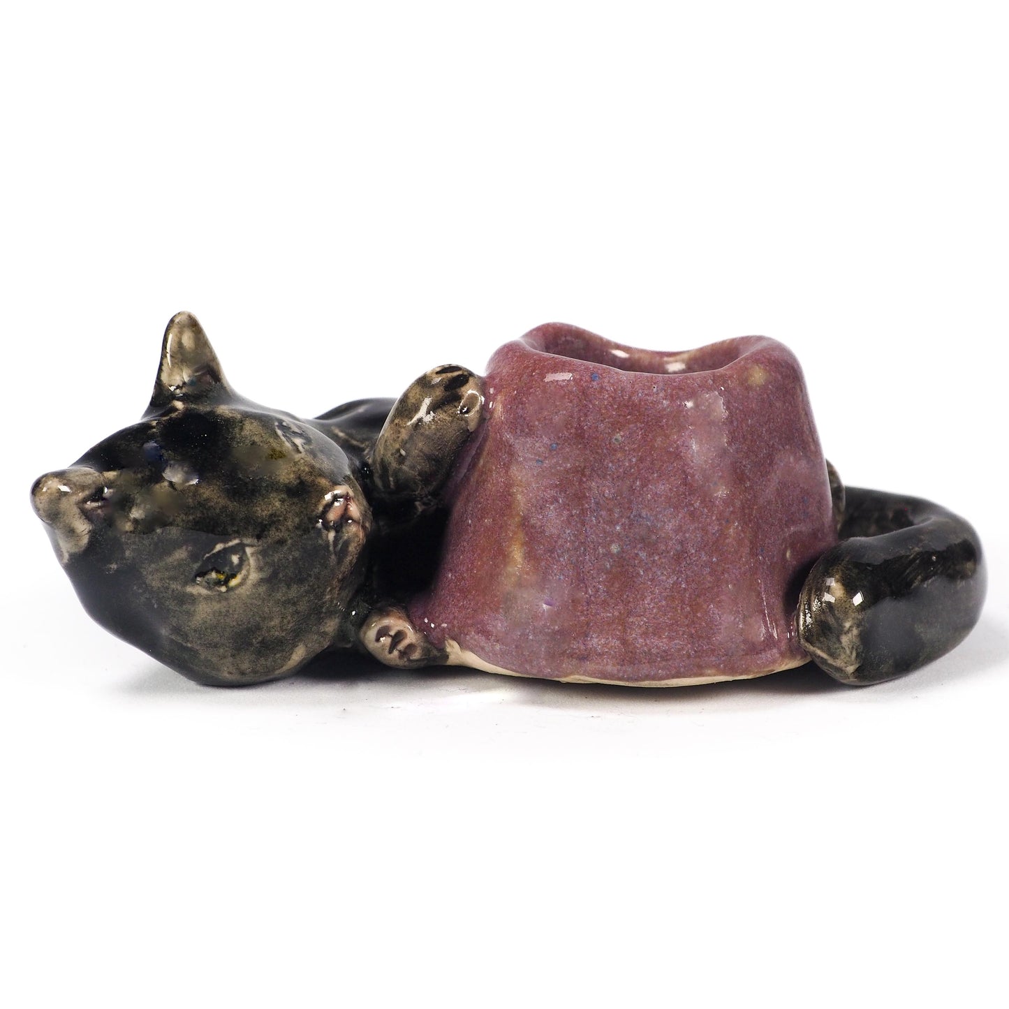 A ceramic cat figurine lying down, holding a small purple bowl or cup in its paws. The cat's head and body are glazed in a dark metallic bronze-black, while the bowl has a textured, speckled purple glaze, by Idania Salcido (Danita Art).