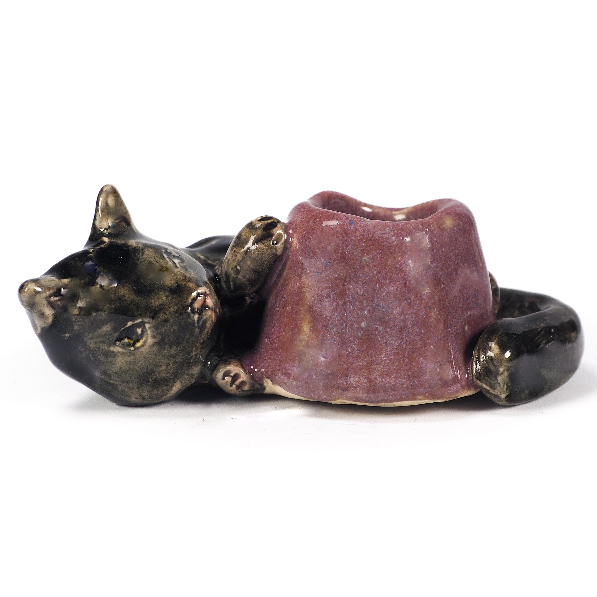 A ceramic cat figurine lying down, holding a small purple bowl or cup in its paws. The cat's head and body are glazed in a dark metallic bronze-black, while the bowl has a textured, speckled purple glaze, by Idania Salcido (Danita Art).