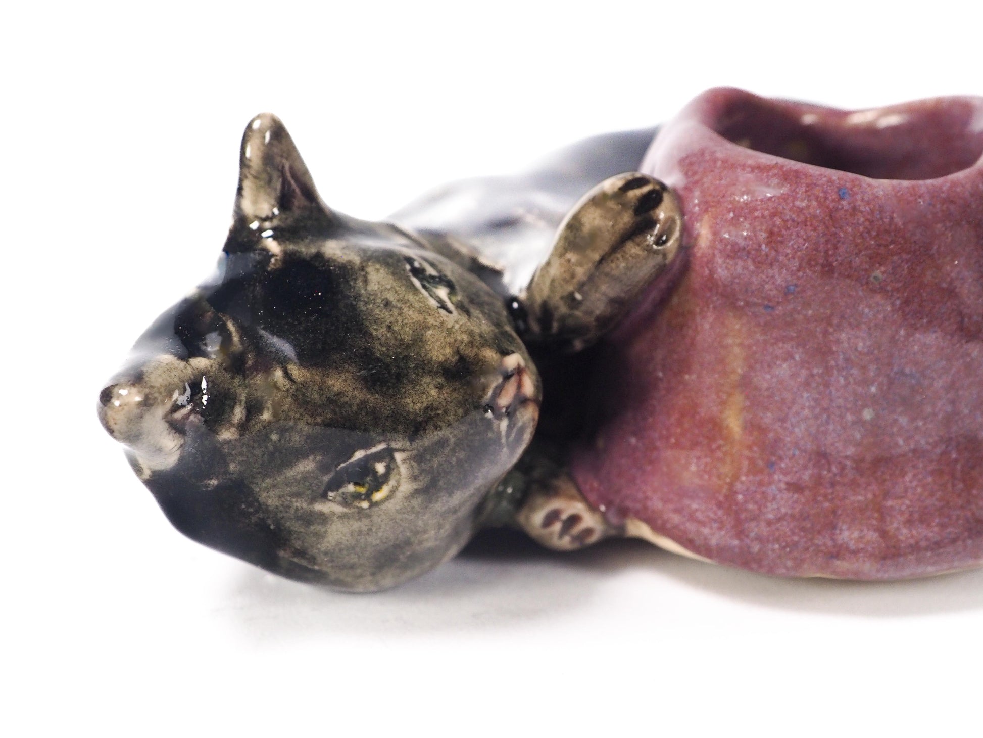 A ceramic cat figurine lying down, holding a small purple bowl or cup in its paws. The cat's head and body are glazed in a dark metallic bronze-black, while the bowl has a textured, speckled purple glaze, by Idania Salcido (Danita Art).