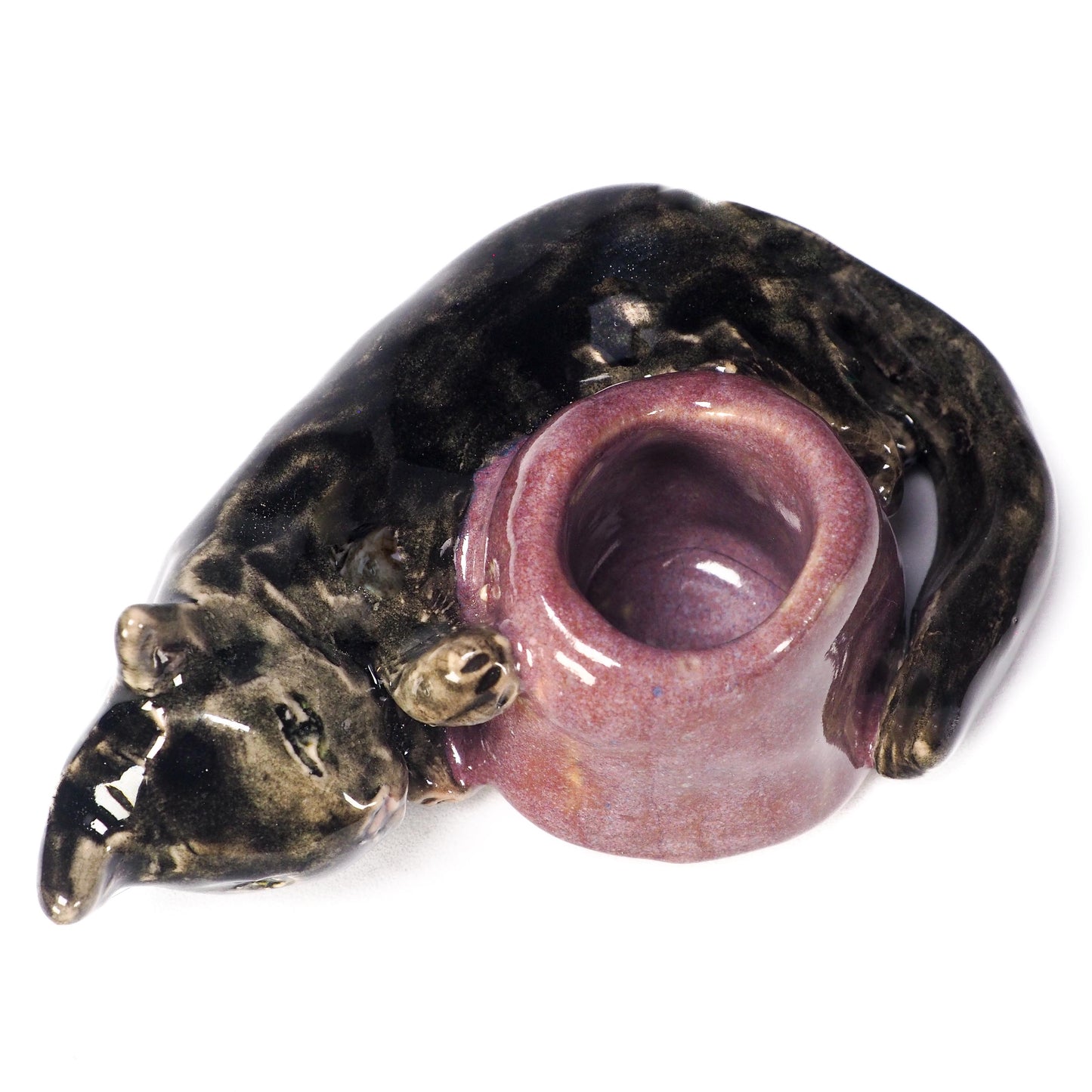A ceramic cat figurine lying down, holding a small purple bowl or cup in its paws. The cat's head and body are glazed in a dark metallic bronze-black, while the bowl has a textured, speckled purple glaze, by Idania Salcido (Danita Art).