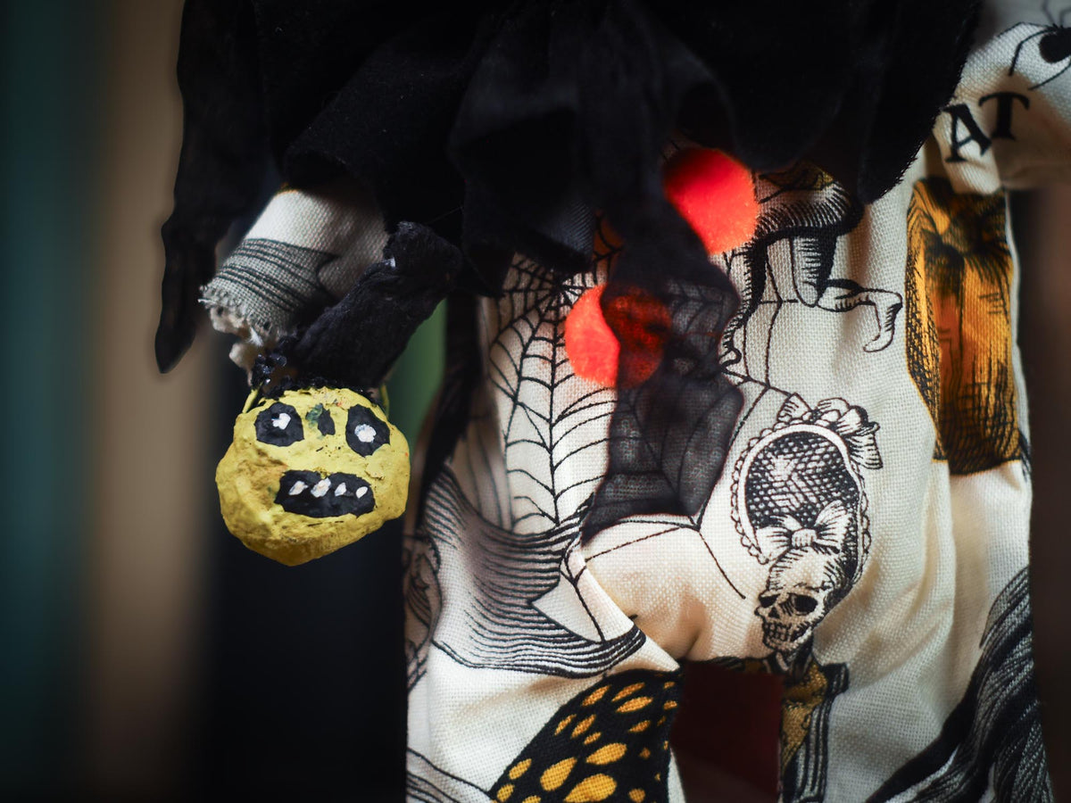 A close-up of a Halloween-themed outfit featuring a black ruffled top, a cream-colored skirt with a spiderweb, skull, and spider print, and a small yellow plush spider accessory held in a black glove.
An original spooky cute outsider folk art Halloween Doll by Idania Salcido (Danita Art). Hand made using spun cotton, paper and fabric to create the perfect Halloween ornament for your tree. Made for someone who appreciates fine and unique art gifts, with lots of love.