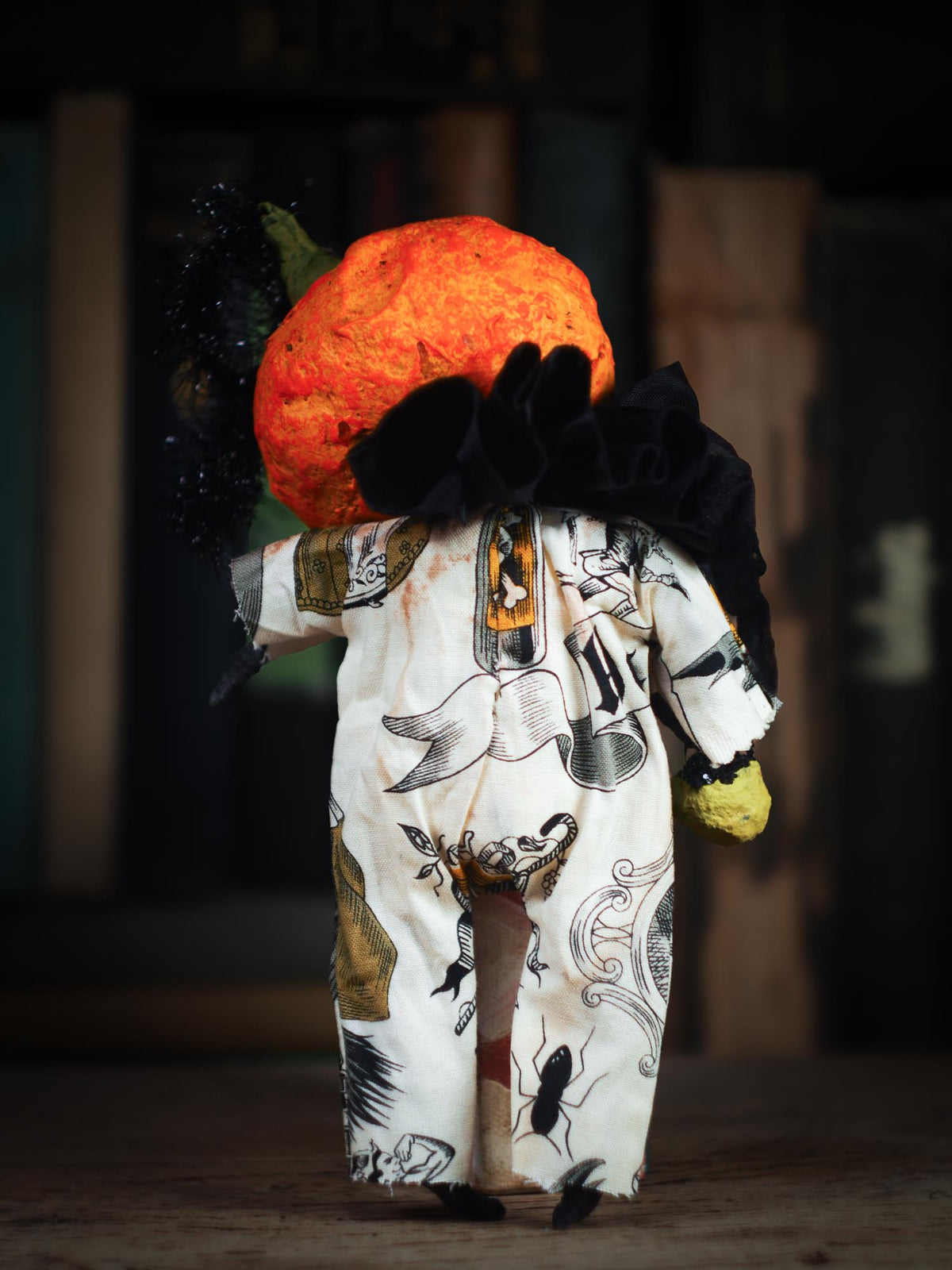 A Halloween-themed doll or figurine shown from the back, wearing a white jumpsuit with black and gold witch illustrations, a large black bow, and an orange pumpkin head against a dark, blurred background.
An original spooky cute outsider folk art Halloween Doll by Idania Salcido (Danita Art). Hand made using spun cotton, paper and fabric to create the perfect Halloween ornament for your tree. Made for someone who appreciates fine and unique art gifts, with lots of love.