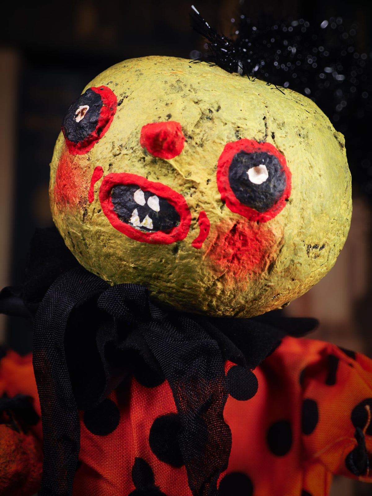 A close-up of a creepy, zombie-like Halloween decoration or doll head with a textured yellow-green face, red and black painted eyes, an open mouth showing teeth, and black tinsel hair, wearing an orange and black polka-dot top. An original spooky cute outsider folk art Halloween Doll by Idania Salcido (Danita Art). Hand made using spun cotton, paper and fabric to create the perfect Halloween ornament for your tree. Made for someone who appreciates fine and unique art gifts, with lots of love.