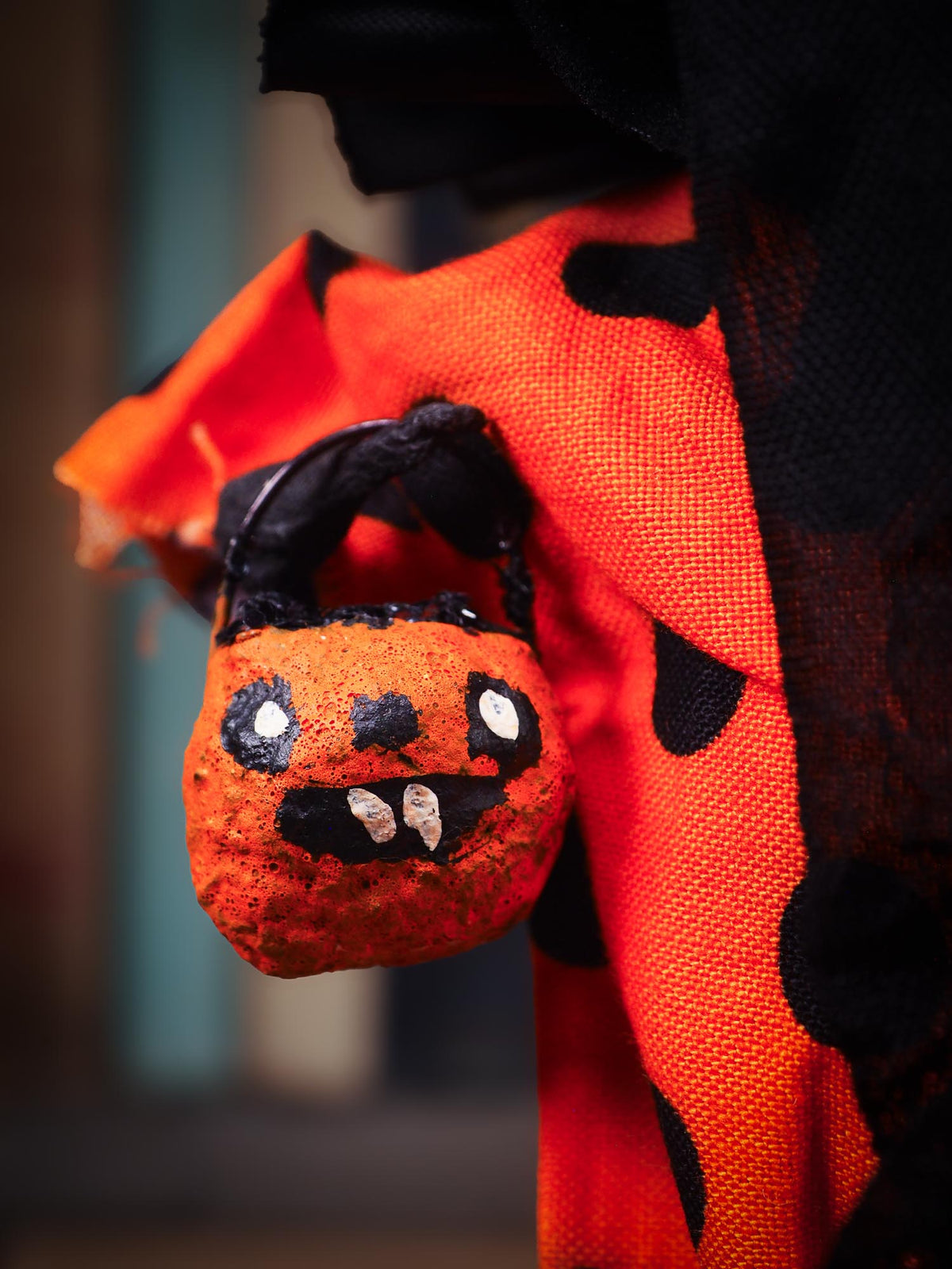 A close-up of a Halloween costume featuring a bright orange fabric with black polka dots and a matching black netted hood, holding a small orange pumpkin-shaped trick-or-treat bucket with a painted spooky face.
An original spooky cute outsider folk art Halloween Doll. Hand made using spun cotton, paper and fabric to create the perfect Halloween ornament for your tree. Specially made for someone who appreciates fine and unique art gifts, a one of a kind creation with an original design by Idania Salcido