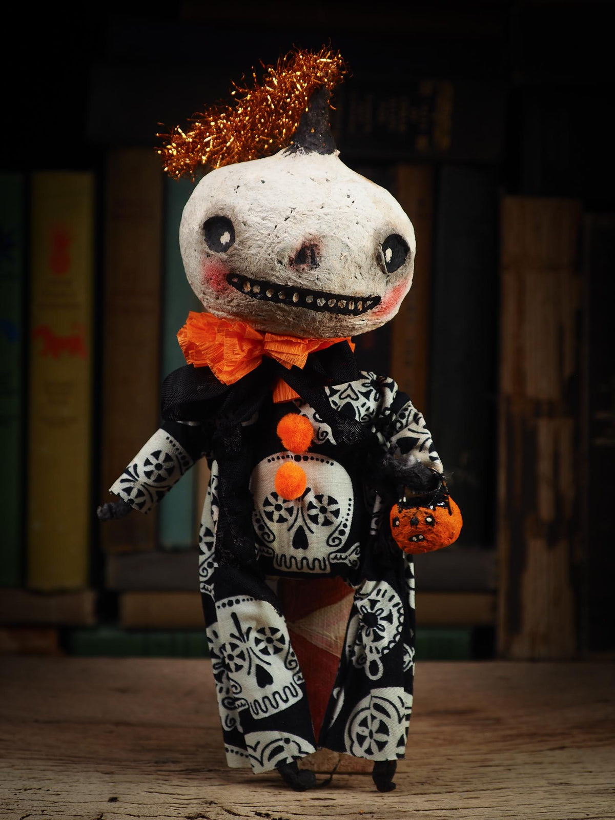 A handmade Halloween doll with a white skull face, black patterned clothing, an orange bow tie, and an orange tinsel hairpiece, standing on a wooden surface against a dark bookshelf background.
An original spooky cute outsider folk art Halloween Doll. Hand made using spun cotton, paper and fabric to create the perfect Halloween ornament for your tree. Specially made for someone who appreciates fine and unique art gifts, It is a one of a kind creation with an original design by Idania Salcido (Danita Art).