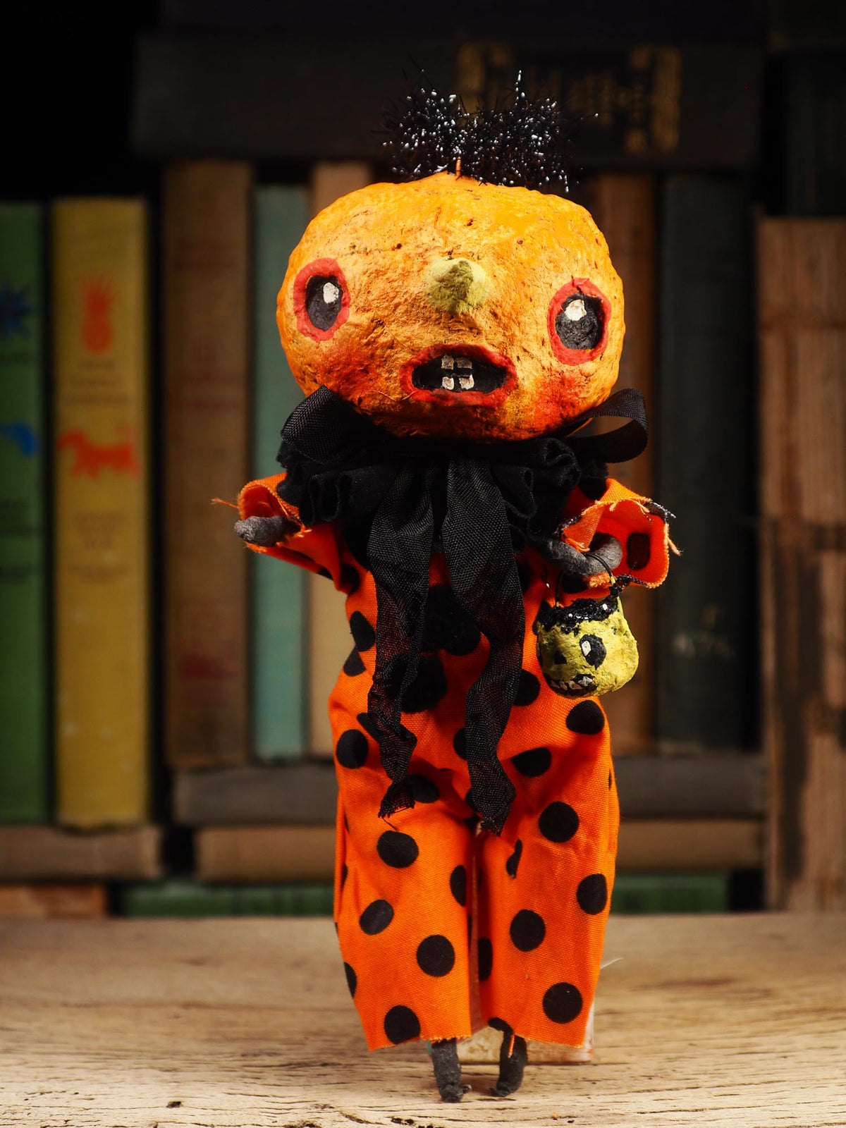 A small handmade doll or figurine with a round orange head, black polka-dot clothing, and a black ruffled collar, standing on a wooden surface in front of a blurred bookshelf background.
An original spooky cute outsider folk art Halloween Doll. Hand made using spun cotton, paper and fabric to create the perfect Halloween ornament for your tree. Specially made for someone who appreciates fine and unique art gifts, It is a one of a kind creation with an original design by Idania Salcido (Danita Art).