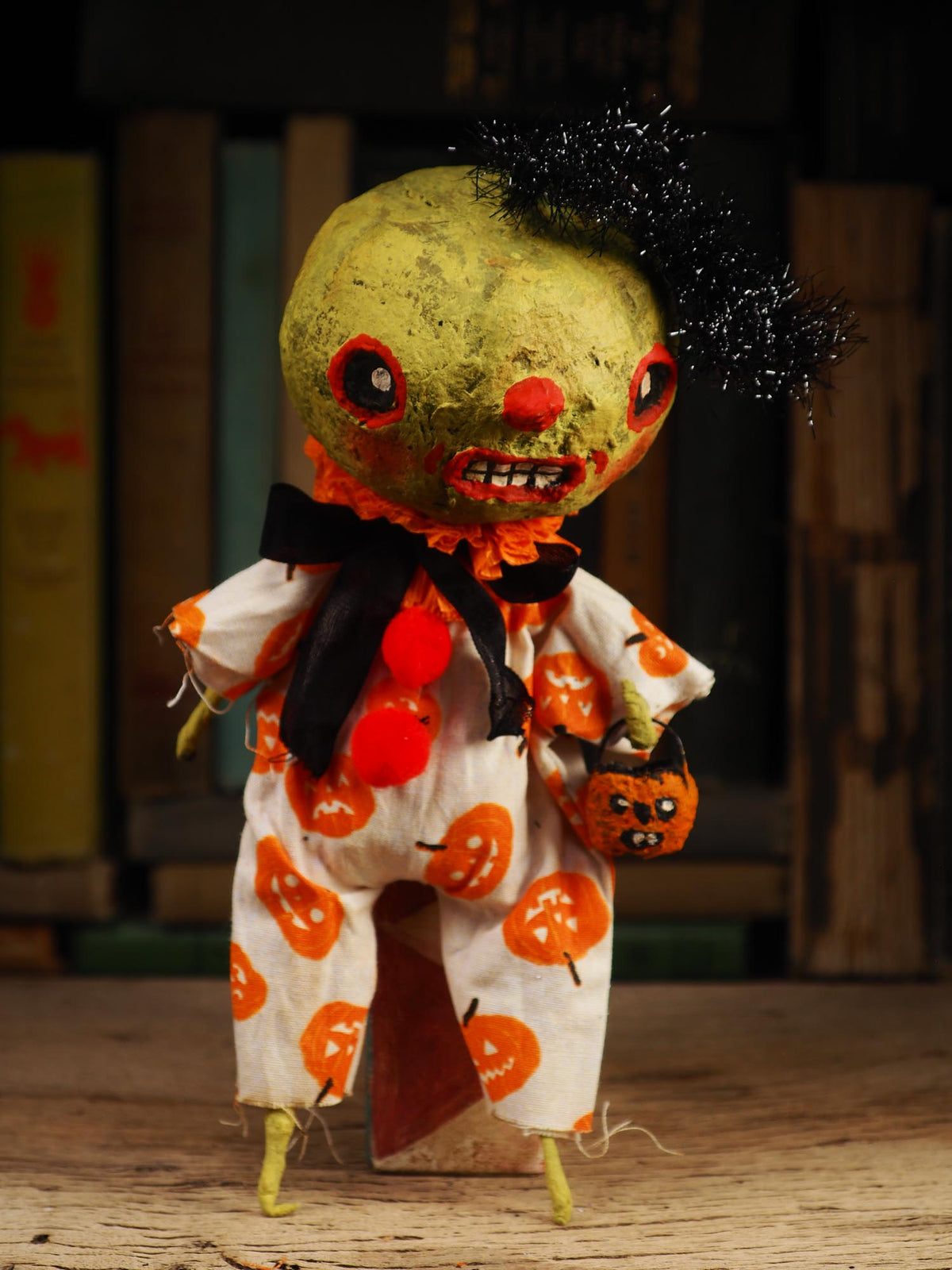 A handmade Halloween doll with a green-yellow face, black tinsel hair, and a white jumpsuit covered in orange pumpkin prints, holding a small jack-o'-lantern bucket.
An original spooky cute outsider folk art Halloween Doll. Hand made using spun cotton, paper and fabric to create the perfect Halloween ornament for your tree. Specially created for someone who appreciates fine and unique art gifts, It is a one of a kind original design by Idania Salcido (Danita Art).