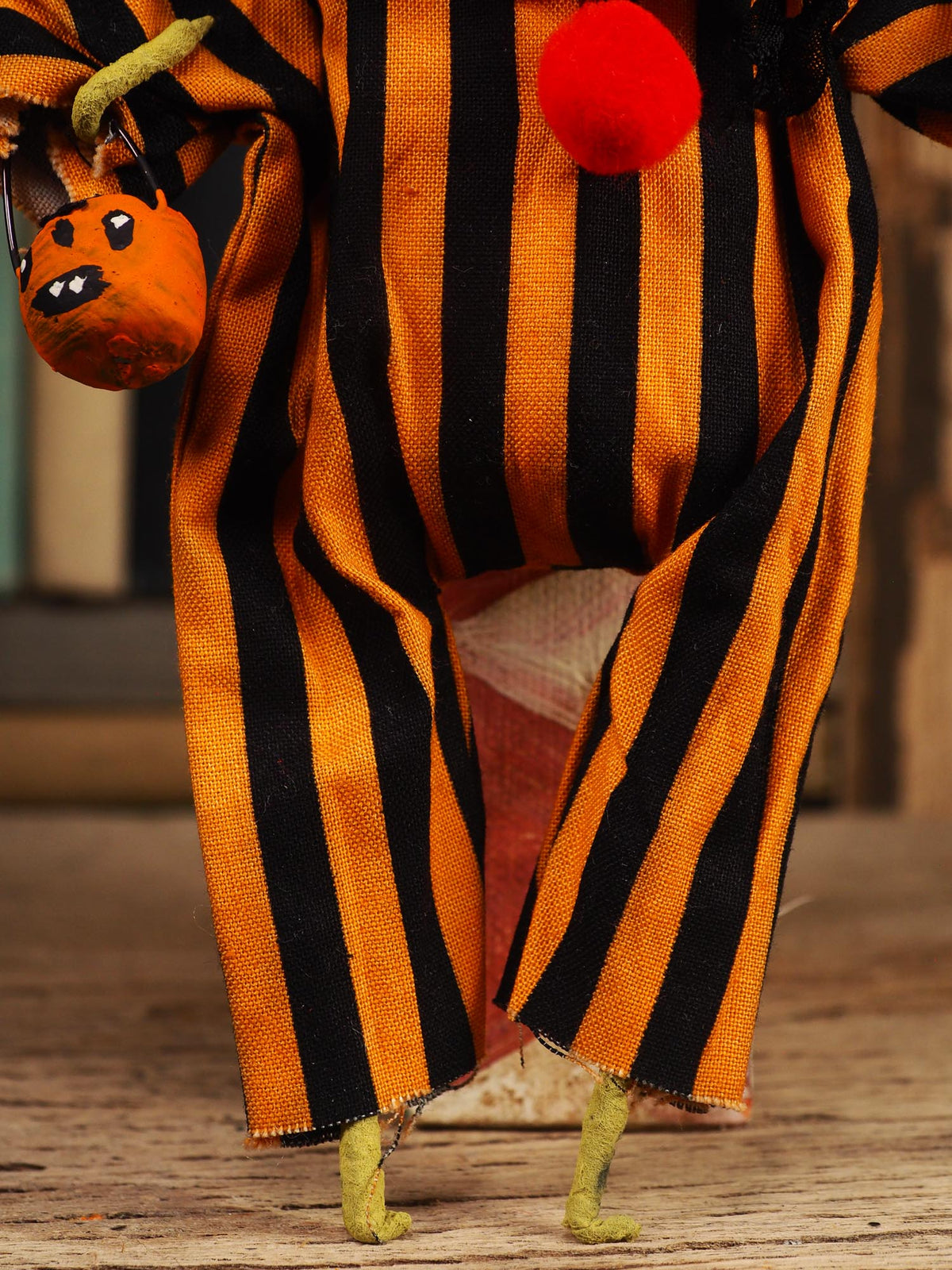 A close-up of a Halloween doll or figurine wearing an orange and black vertically striped costume, holding a small orange pumpkin bag, with a red pom-pom on the chest and green hands and feet.
An original spooky cute outsider folk art Halloween Doll. Hand made using spun cotton, paper and fabric to create the perfect Halloween ornament for your tree. Specially created for someone who appreciates fine and unique art gifts, It is a one of a kind original design by Idania Salcido (Danita Art).