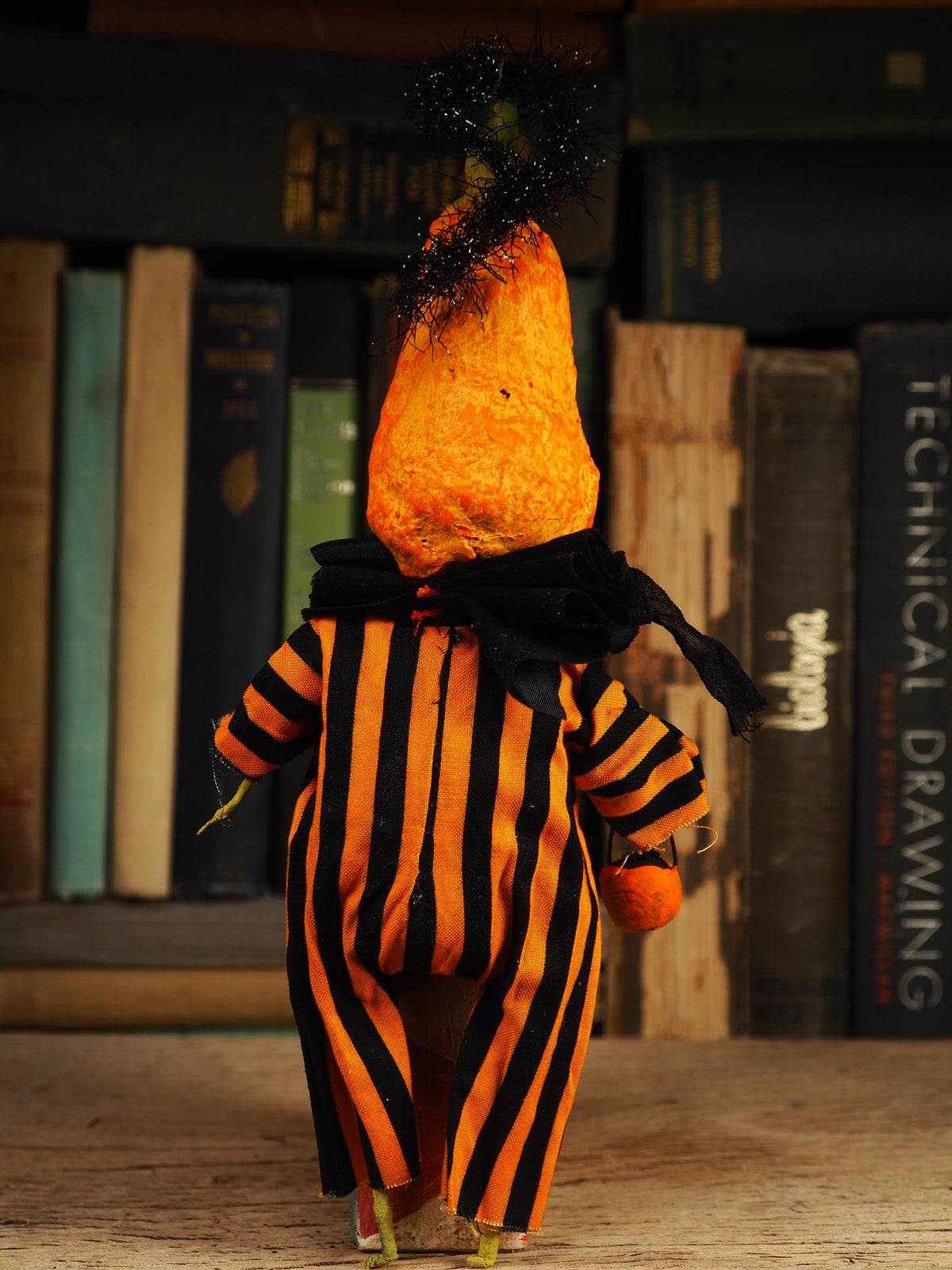 A small Halloween-style figurine or doll shown from the back, wearing an orange-and-black vertically striped outfit with a large orange cone-shaped head and black tinsel on top, standing in front of a bookshelf.
An original spooky cute outsider folk art Halloween Doll. Hand made using spun cotton, paper and fabric to create the perfect Halloween ornament for your tree. Specially created for someone who appreciates fine and unique art gifts, It is a one of a kind original design by Idania Salcido (Danita Art