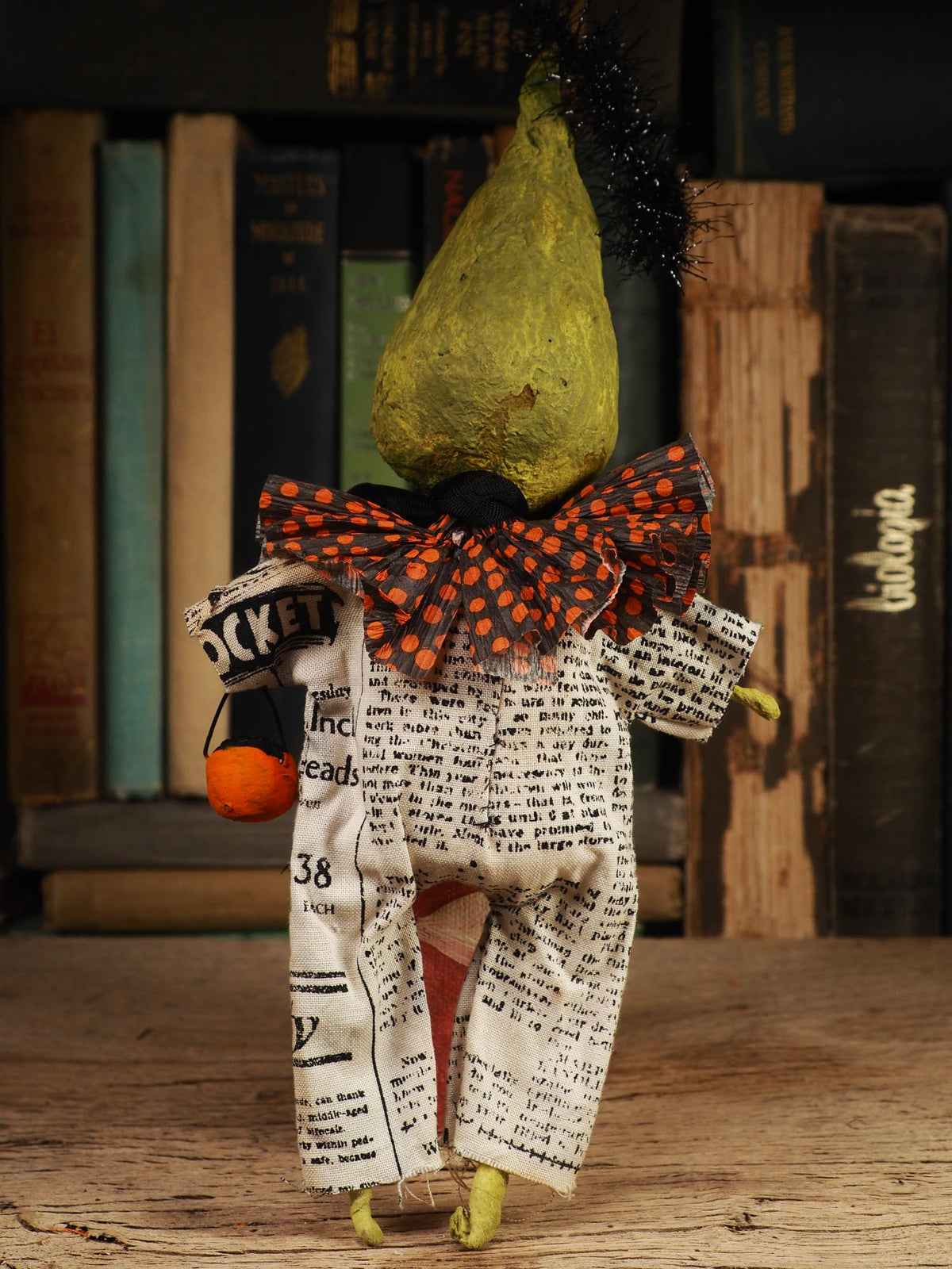 A handmade Halloween doll with a green pear-shaped head, black tinsel hair, a red and black polka-dot bow, and newspaper-print pants, posed on a wooden surface in front of a bookshelf.
An original spooky cute outsider folk art Halloween Doll. Hand made using spun cotton, paper and fabric to create the perfect Halloween ornament for your tree. Specially created for someone who appreciates fine and unique art gifts, It is a one of a kind original design by Idania Salcido (Danita Art).