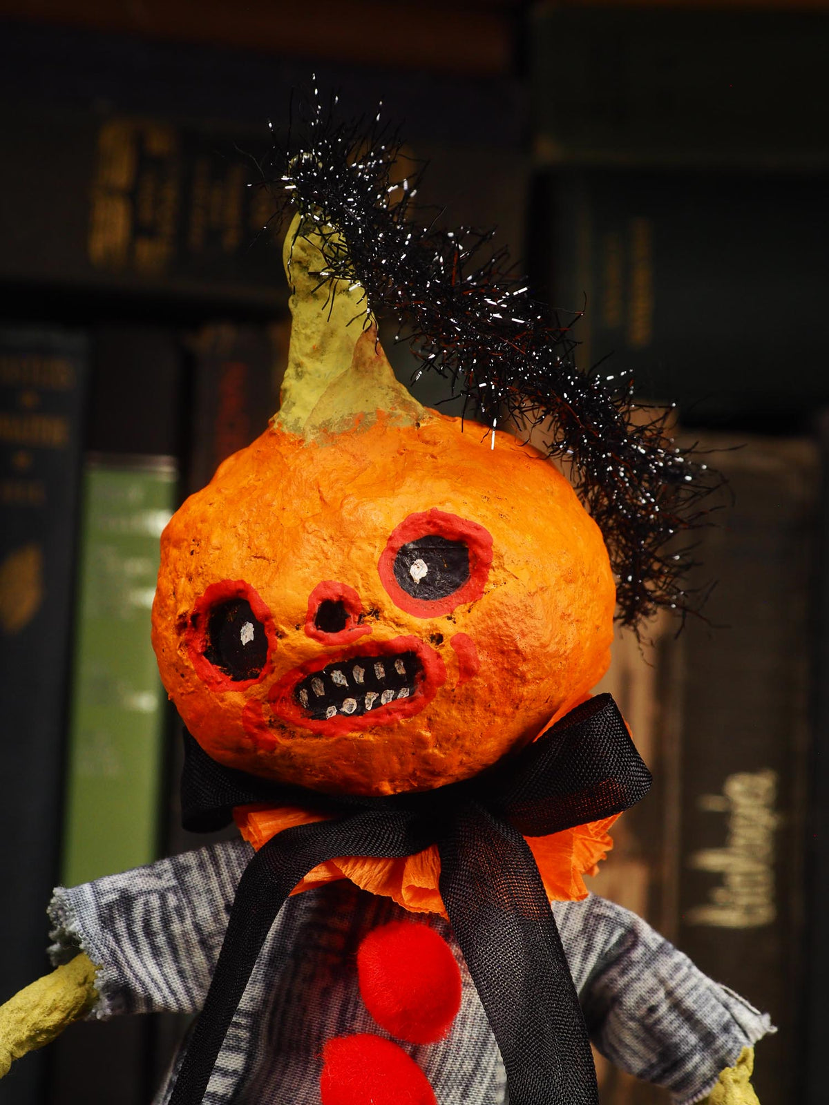 A creepy Halloween pumpkin doll with a textured orange head, black tinsel hair, a black ribbon bow, and a striped shirt, set against a dark background of bookshelves.
An original handmade spun cotton Halloween art doll by Idania Salcido (Danita Art).