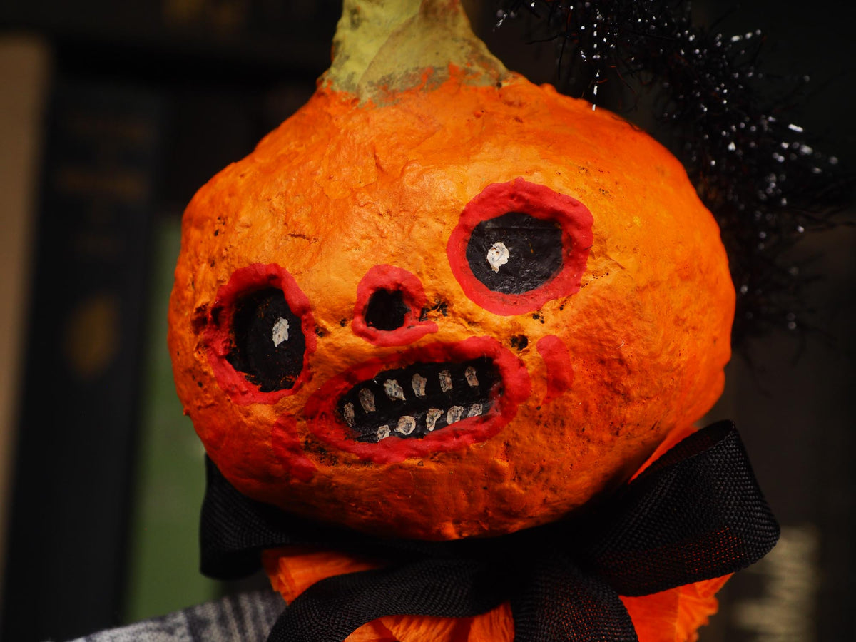 A close-up of a Halloween pumpkin decoration with a distressed orange surface, black eye and mouth details, a black ribbon bow at the neck, and black tinsel on top, photographed against a dark blurred background.
An original handmade spun cotton Halloween art doll by Idania Salcido (Danita Art).