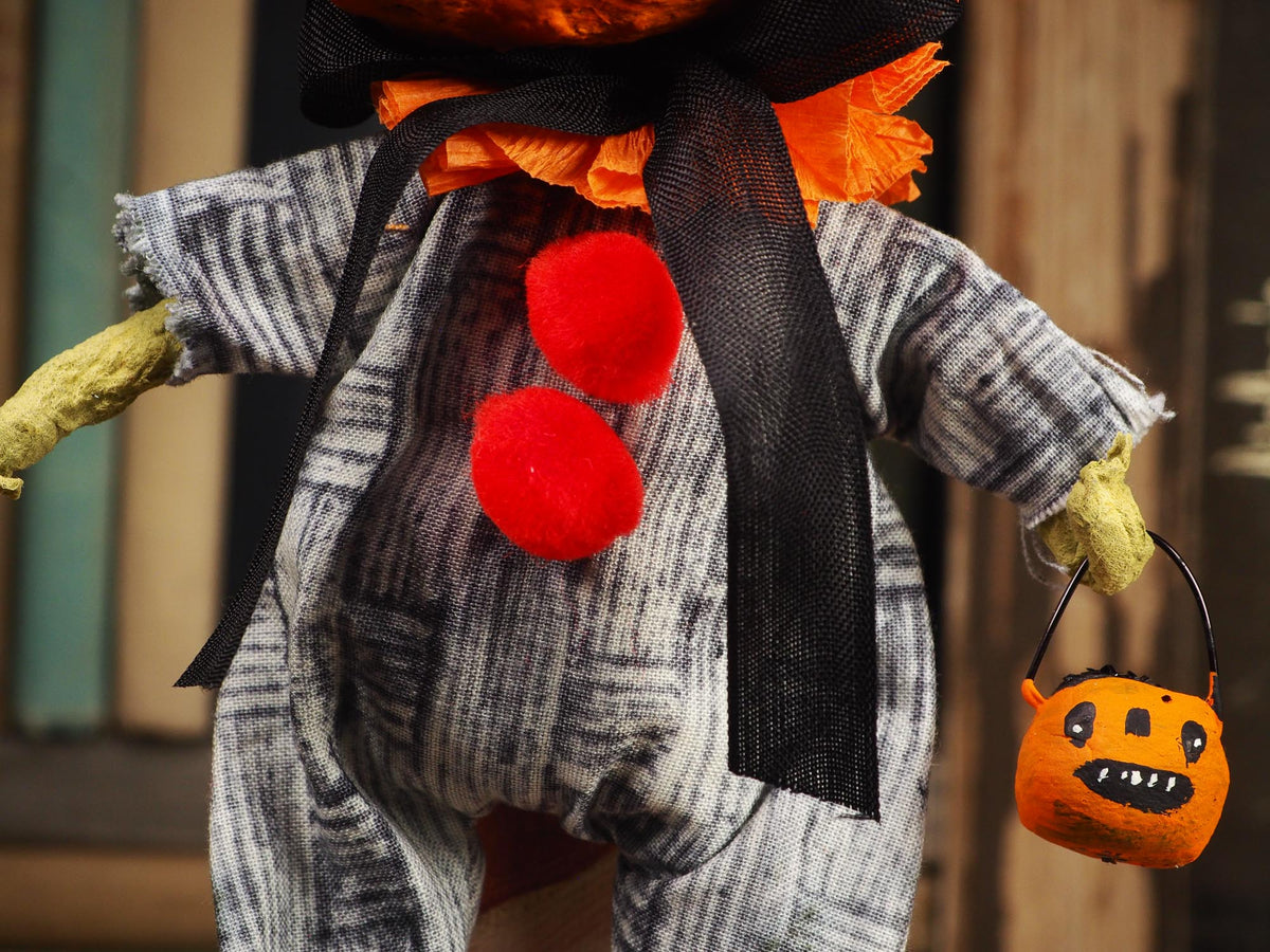 A Halloween scarecrow doll wearing a gray and white striped shirt, black mesh bow, orange flower headpiece, and holding an orange pumpkin bucket against a blurred rustic background.
An original handmade spun cotton Halloween art doll by Idania Salcido (Danita Art).