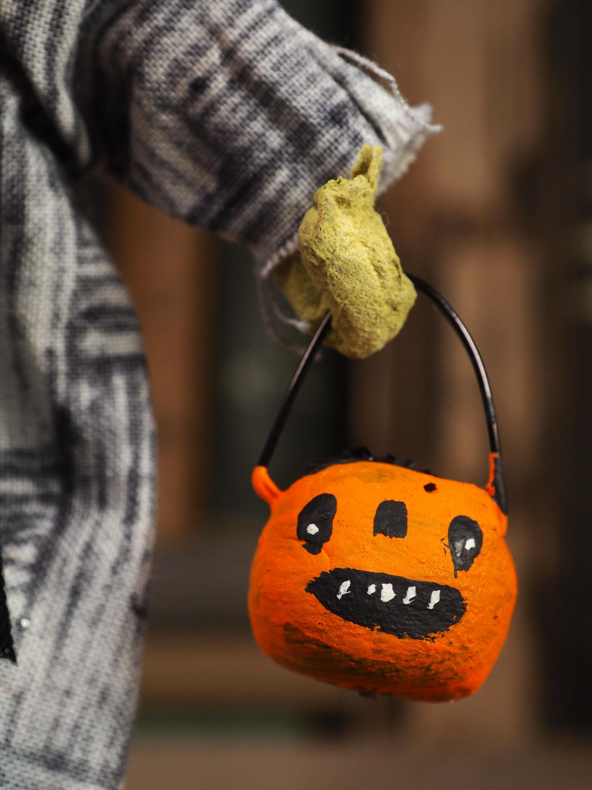 A person wearing a gray plaid sweater and a yellow-green work glove holds a small orange jack-o'-lantern bucket with a black handle against a blurred outdoor background.
An original handmade spun cotton Halloween art doll by Idania Salcido (Danita Art).
