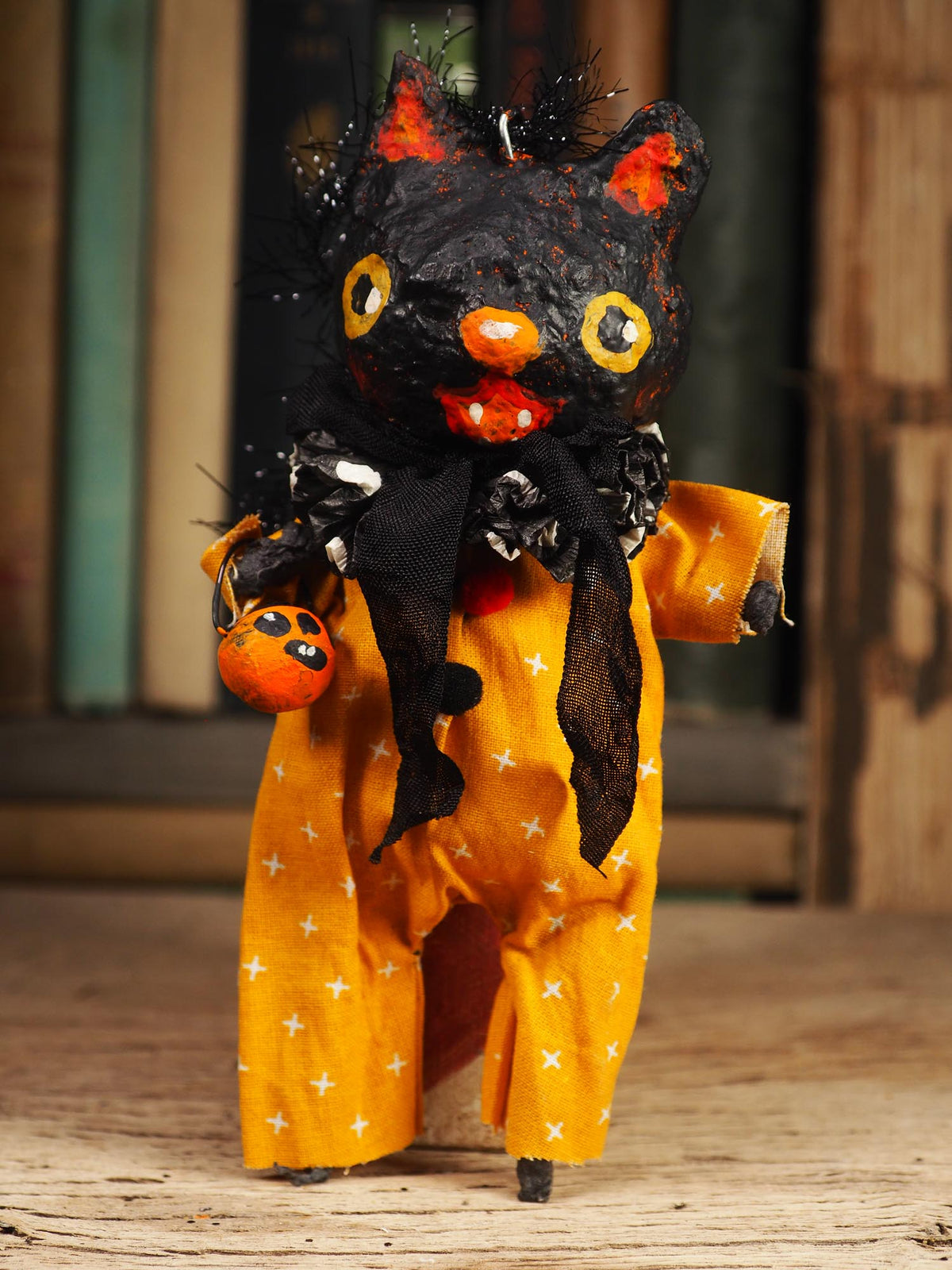 A handmade Halloween cat doll with a black textured body, orange nose and mouth, yellow eyes, and an orange jumpsuit with white stars, holding a small pumpkin on a wooden surface.
An original handmade spun cotton Halloween art doll by Idania Salcido (Danita Art).