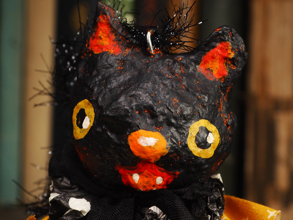 A close-up of a black cat-shaped decorative object with orange and yellow painted details, black tinsel on its head, and a black fabric base, photographed against a blurred indoor background.
An original handmade spun cotton Halloween art doll by Idania Salcido (Danita Art).