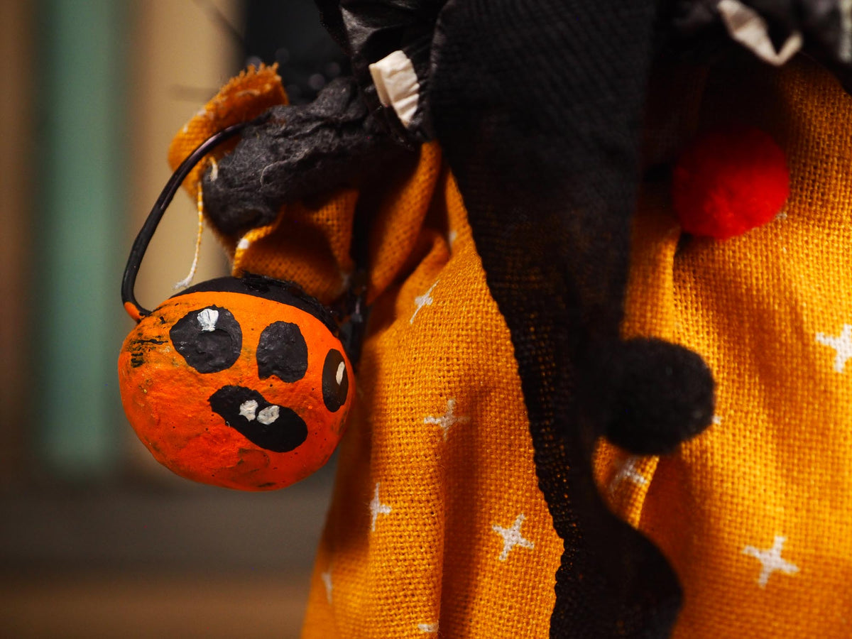 A close-up of a Halloween costume featuring a textured orange dress with white star patterns, a black pointed hat with black pom-poms, and a small orange pumpkin-shaped bucket accessory with a black handle.
An original handmade spun cotton Halloween art doll by Idania Salcido (Danita Art).