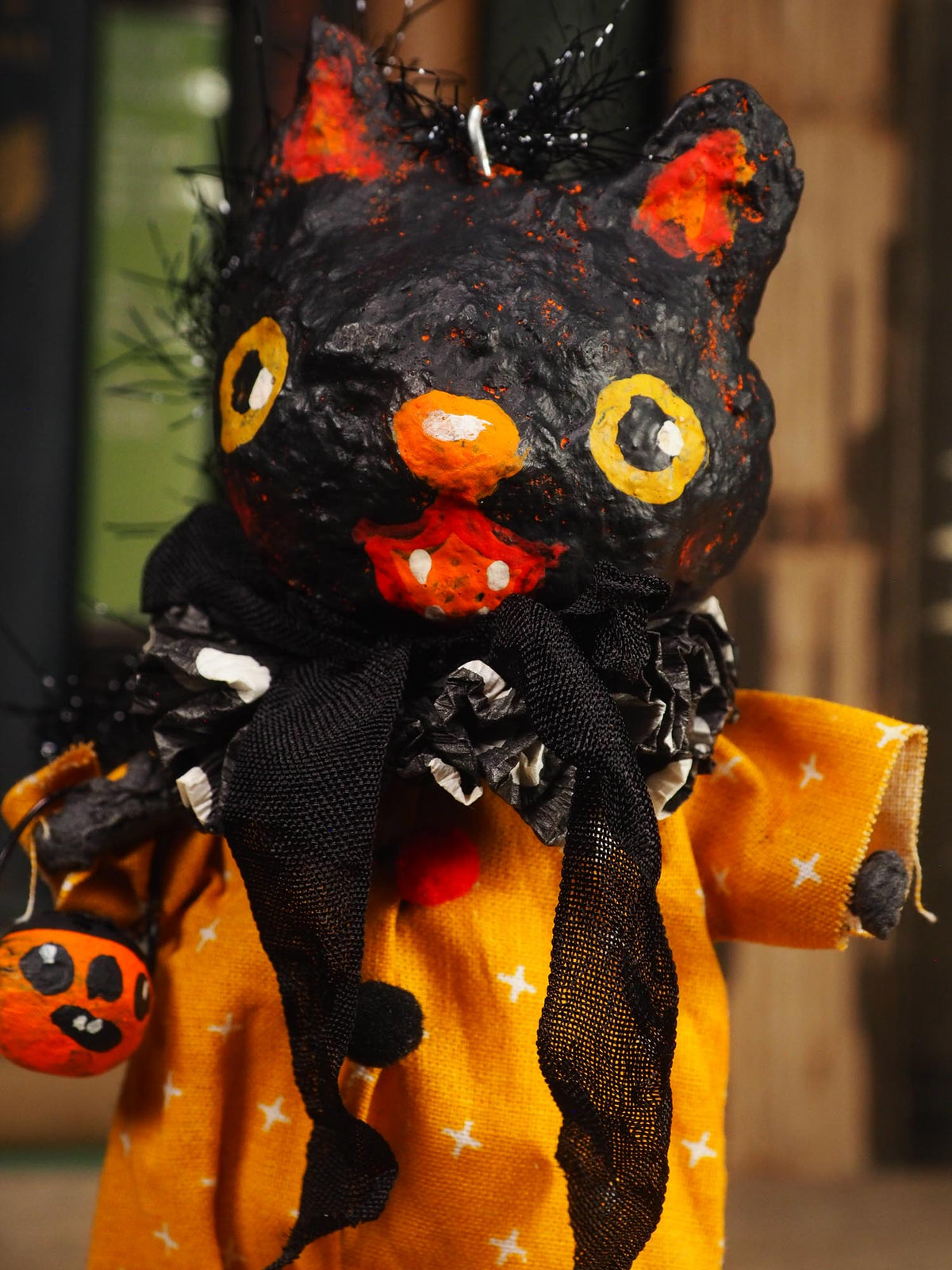 A handmade Halloween cat doll with a black textured hat, orange and yellow painted face, black ruffled collar, and an orange dress with white stars, holding a small pumpkin.
An original handmade spun cotton Halloween art doll by Idania Salcido (Danita Art).