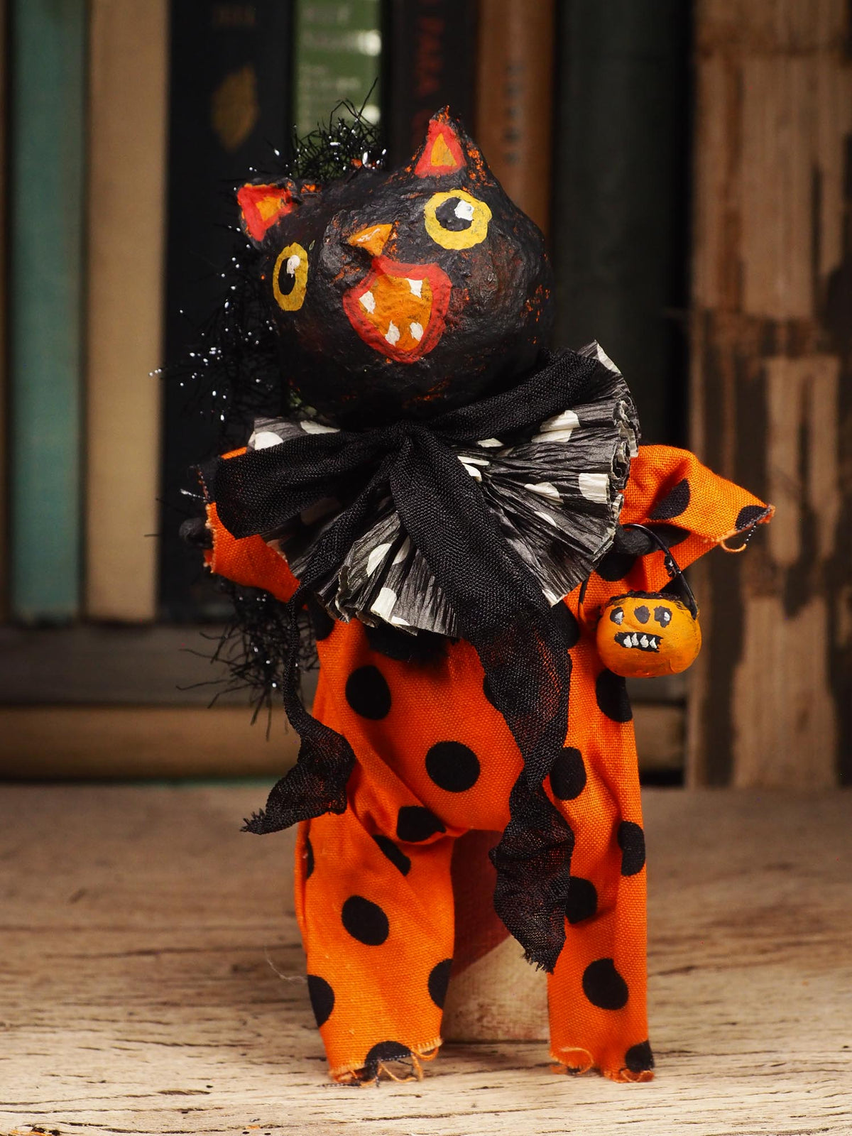 A handmade Halloween doll or figurine with a black cat head, yellow eyes, and an orange face, wearing an orange and black polka-dot body and a black-and-white ruffled collar, standing on a wooden surface.
An original handmade spun cotton Halloween art doll by Idania Salcido (Danita Art).