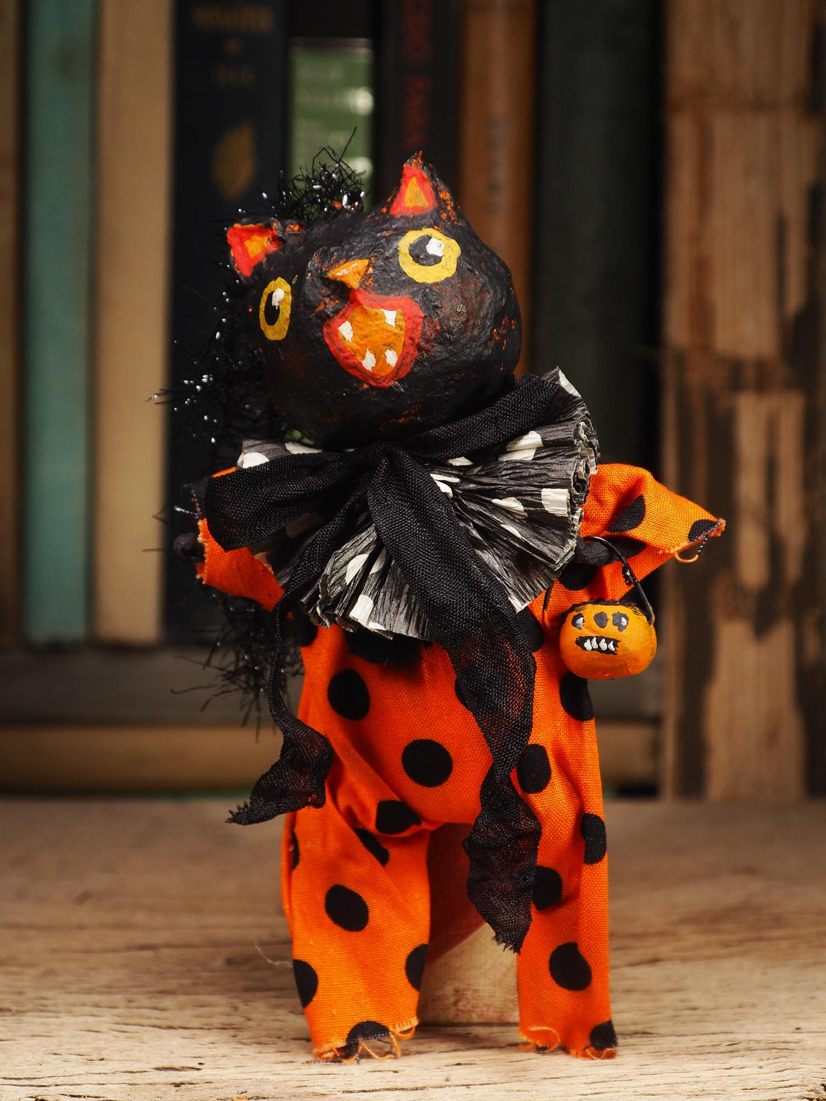 A handmade Halloween cat figurine with a black body, orange polka-dotted dress, black-and-white checkered bow, and a small pumpkin bag, standing on a wooden surface.
An original handmade spun cotton Halloween art doll by Idania Salcido (Danita Art).