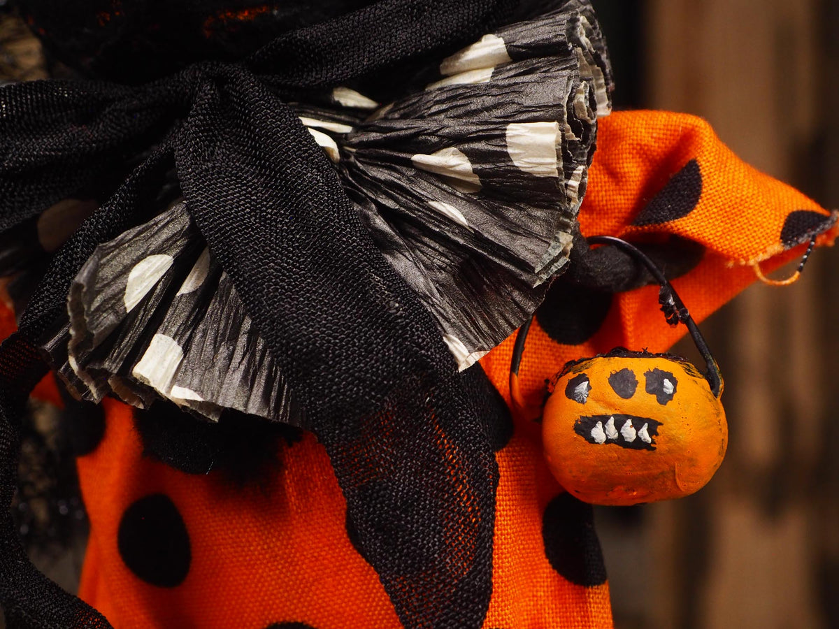 Close-up of a Halloween decoration featuring an orange fabric with black polka dots, a large black tulle bow with white polka dots, and a small orange pumpkin bucket ornament with a painted scary face.
An original handmade spun cotton Halloween art doll by Idania Salcido (Danita Art).