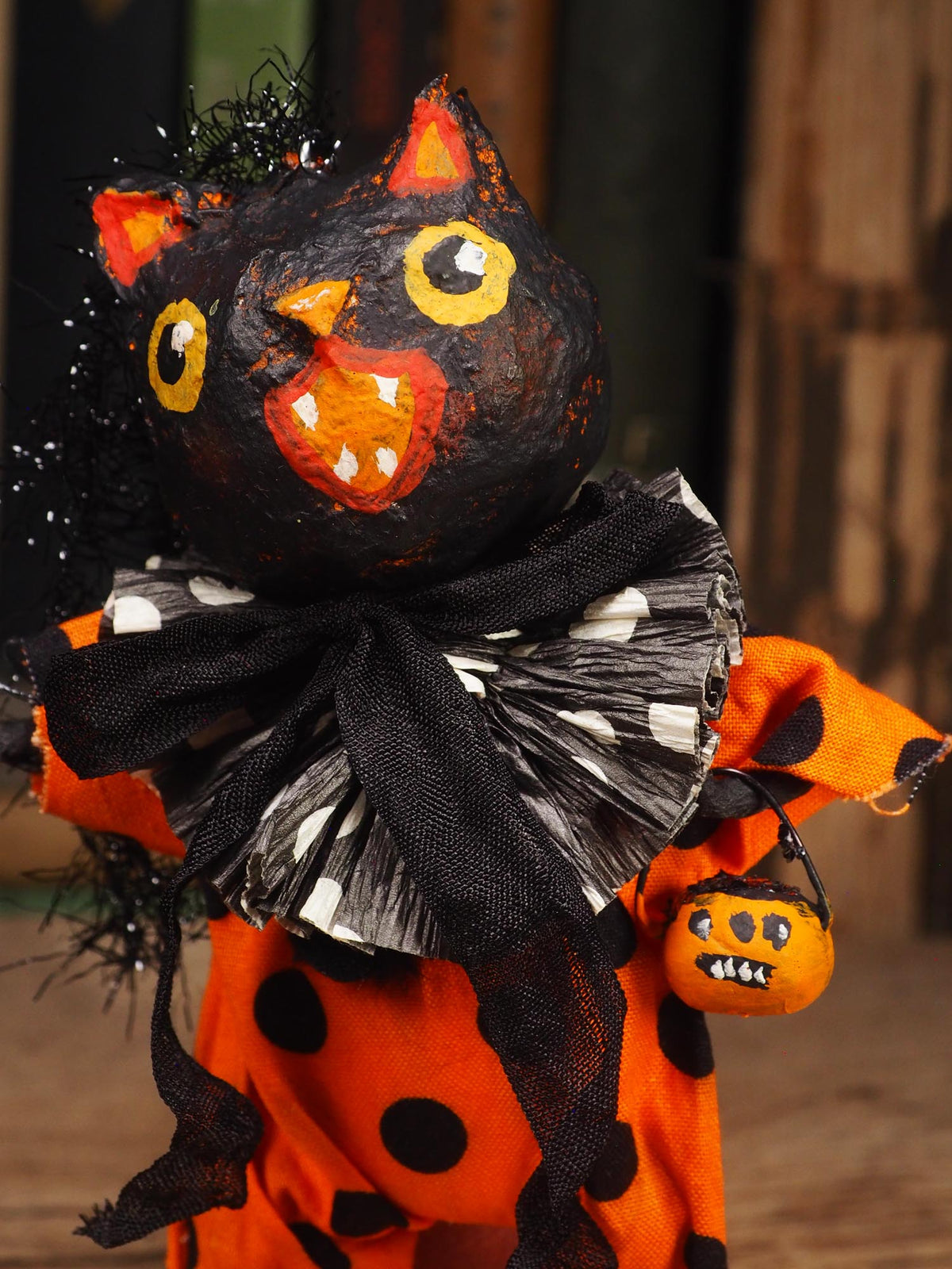 A Halloween decorative figure with a black cat-like head, orange body, black polka dots, a black-and-white ruffled collar, and a small orange pumpkin bucket, photographed against a blurred rustic background.
An original handmade spun cotton Halloween art doll by Idania Salcido (Danita Art).