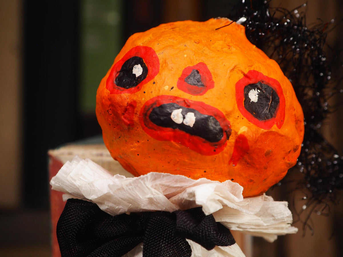 An orange Halloween decoration shaped like a pumpkin with a scary painted face, black tinsel hair, a white ruffled collar, and a black bow, photographed close up against a dark blurred background.
An original handmade spun cotton Halloween art doll by Idania Salcido (Danita Art).