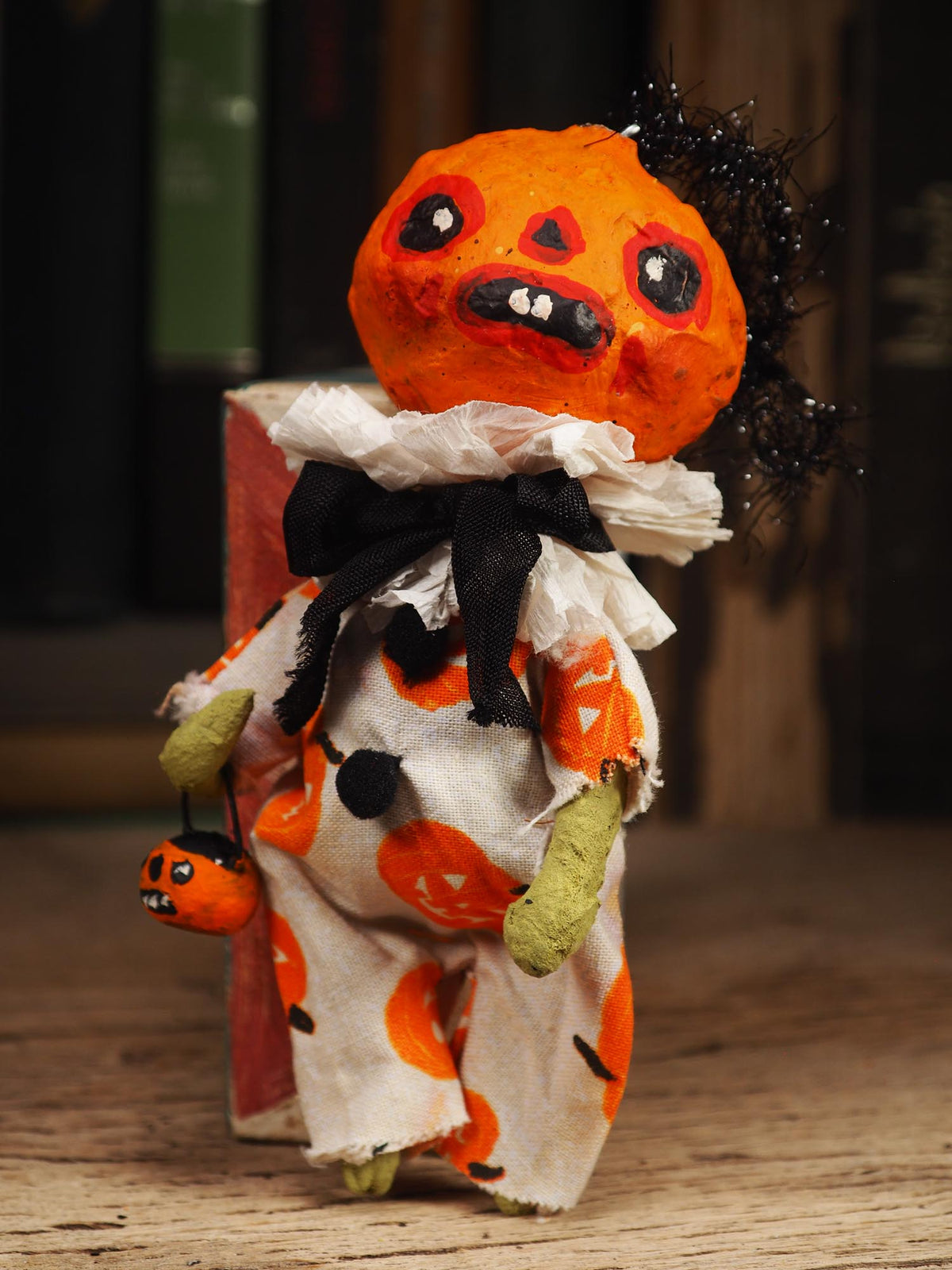 A handmade Halloween doll with a painted orange pumpkin head, black hair, and a tattered white dress with orange pumpkin prints, standing on a wooden surface.
An original handmade spun cotton Halloween art doll by Idania Salcido (Danita Art).
