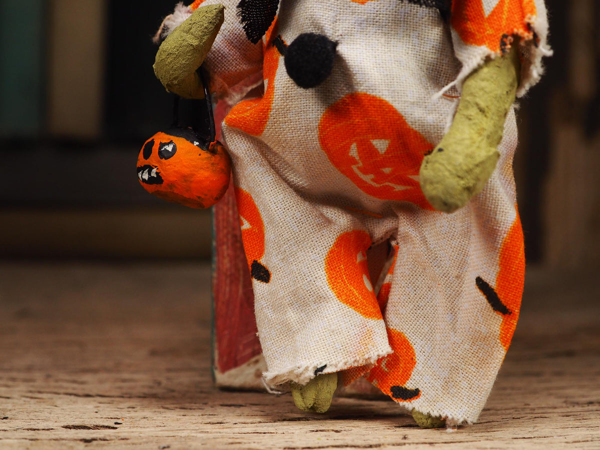 A close-up of a Halloween-themed rag doll or toy figure wearing a tattered off-white dress with orange jack-o'-lantern prints, olive green rag limbs, and holding an orange pumpkin bag.
An original handmade spun cotton Halloween art doll by Idania Salcido (Danita Art).
