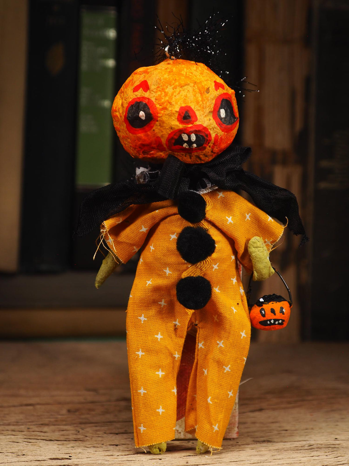 A handmade Halloween doll with a stitched orange pumpkin head, black eyes, and a black collar, wearing an orange jumpsuit with white star patterns and holding a small orange jack-o'-lantern bucket. Original spun cotton hand made Halloween doll ornament by Idania Salcido (Danita Art)