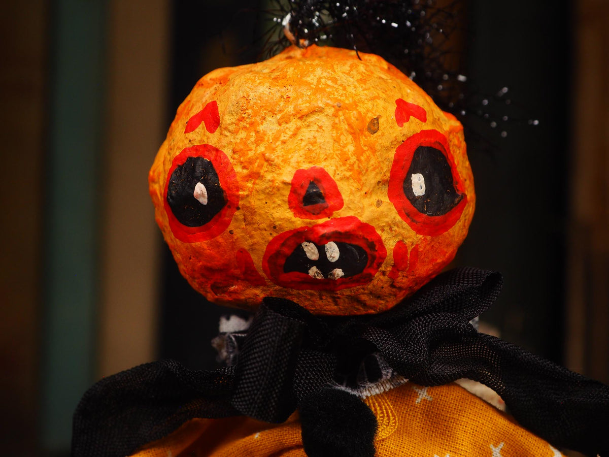 A close-up of a Halloween decorative figure with a textured orange pumpkin head, hand-painted red and black facial features, black fabric around the neck, and an orange garment below. Original spun cotton hand made Halloween doll ornament by Idania Salcido (Danita Art)