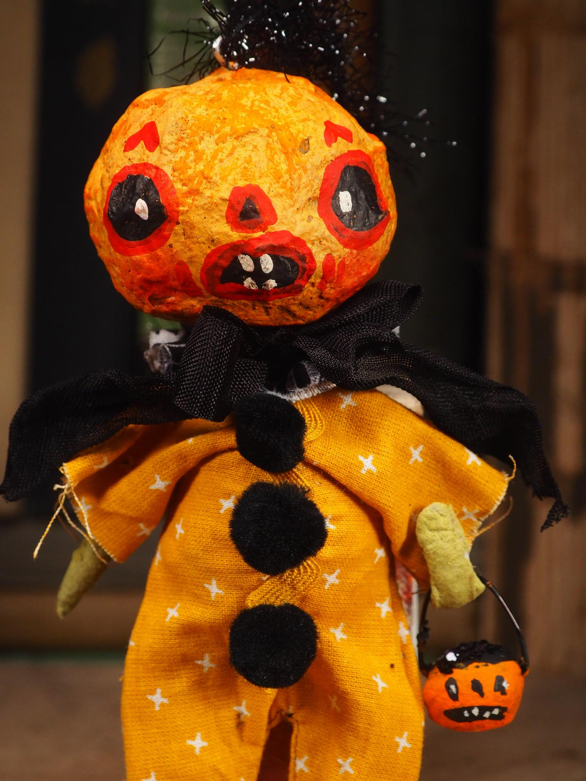 A Halloween-style rag doll or decorative figure with a distressed orange pumpkin head, black hair, a black cape, and a yellow outfit with white star prints, holding a small orange pumpkin bucket against a dark blurred background. Original spun cotton hand made Halloween doll ornament by Idania Salcido (Danita Art)