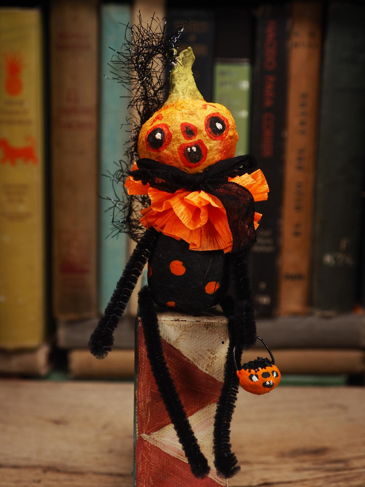 A handmade Halloween decoration shaped like a gourd-headed creature with large black eyes, an orange and black ruffled collar, black pipe-cleaner limbs, and a small orange pumpkin bucket, set against a blurred background of book spines. Original spun cotton hand made Halloween doll ornament by Idania Salcido (Danita Art)