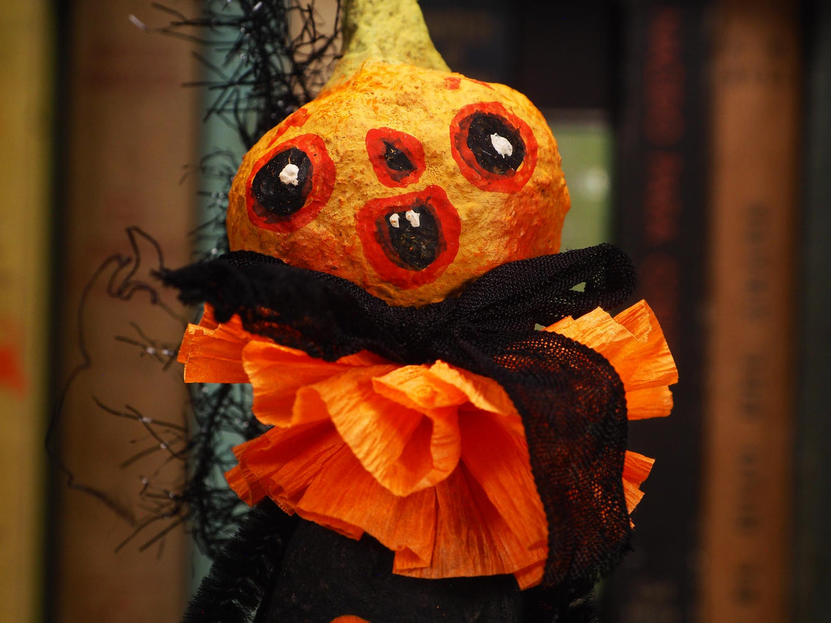 A Halloween pumpkin decoration with a painted spooky face, black tinsel accents, and an orange-and-black ruffled collar, photographed against a blurred background of book spines. Original spun cotton hand made Halloween doll ornament by Idania Salcido (Danita Art)