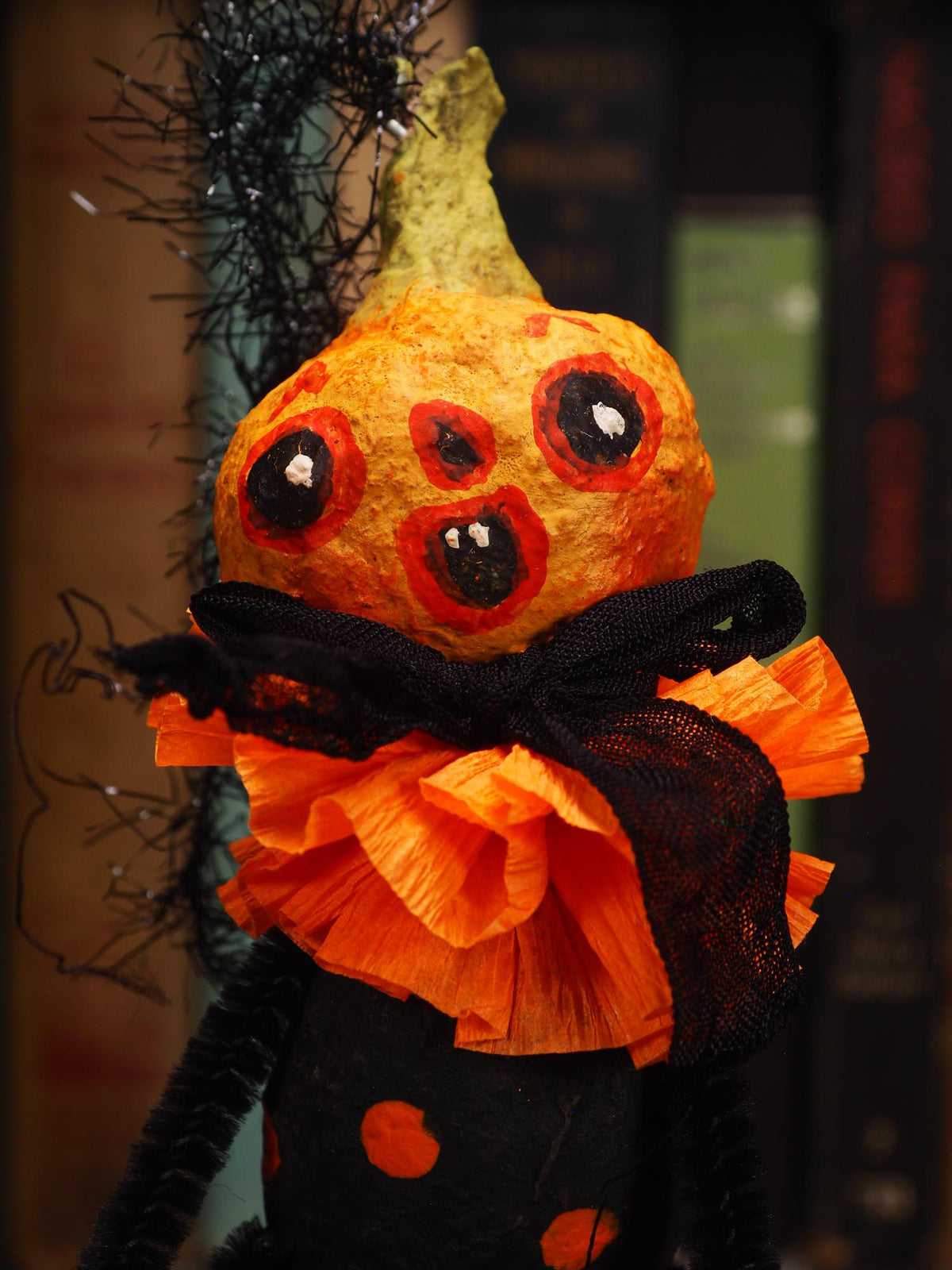A Halloween decoration shaped like a small orange pumpkin with three large painted eyes and an open mouth, wearing an orange ruffled collar and black mesh ribbon, with black pipe cleaners extending from the sides. Original spun cotton hand made Halloween doll ornament by Idania Salcido (Danita Art)