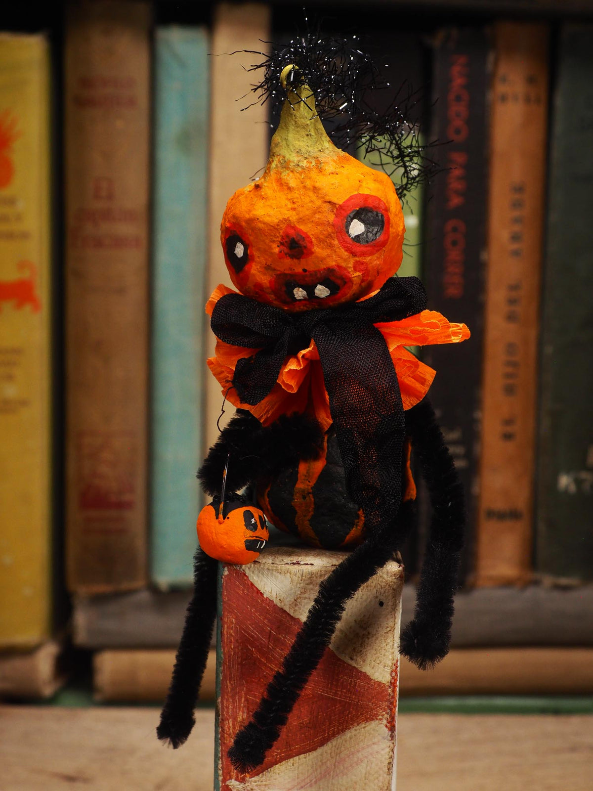 A handmade Halloween decoration shaped like a pumpkin-headed spider, featuring black pipe cleaners for legs, a black bow, and orange and black striped body details, posed in front of a blurred background of book spines. Original spun cotton hand made Halloween doll ornament by Idania Salcido (Danita Art)