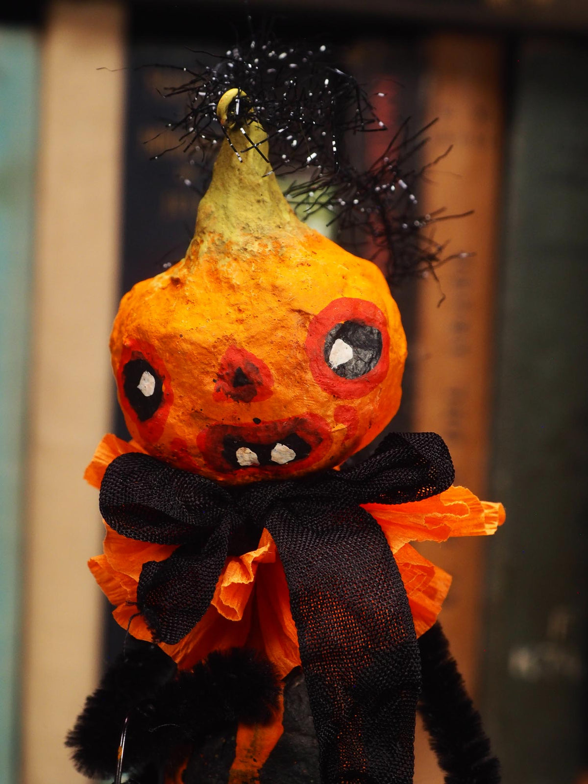 A handmade Halloween decoration shaped like a distorted orange gourd with a green top, black tinsel on the head, a black ribbon bow around the neck, and orange-and-black ruffled fabric at the base, photographed against a blurred indoor background. Original spun cotton hand made Halloween doll by Idania Salcido (Danita Art)