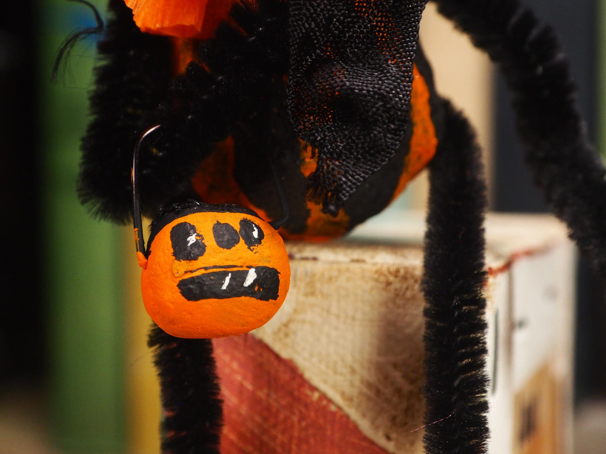 A close-up of a Halloween spider decoration with black pipe cleaner legs, orange fabric body sections, and a small orange pumpkin bucket hanging from a hook. Original spun cotton hand made Halloween doll by Idania Salcido (Danita Art)