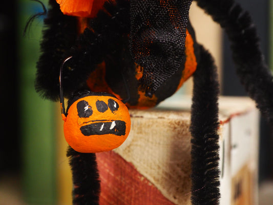 A close-up of a Halloween spider decoration with black pipe cleaner legs, orange fabric body sections, and a small orange pumpkin bucket hanging from a hook. Original spun cotton hand made Halloween doll by Idania Salcido (Danita Art)