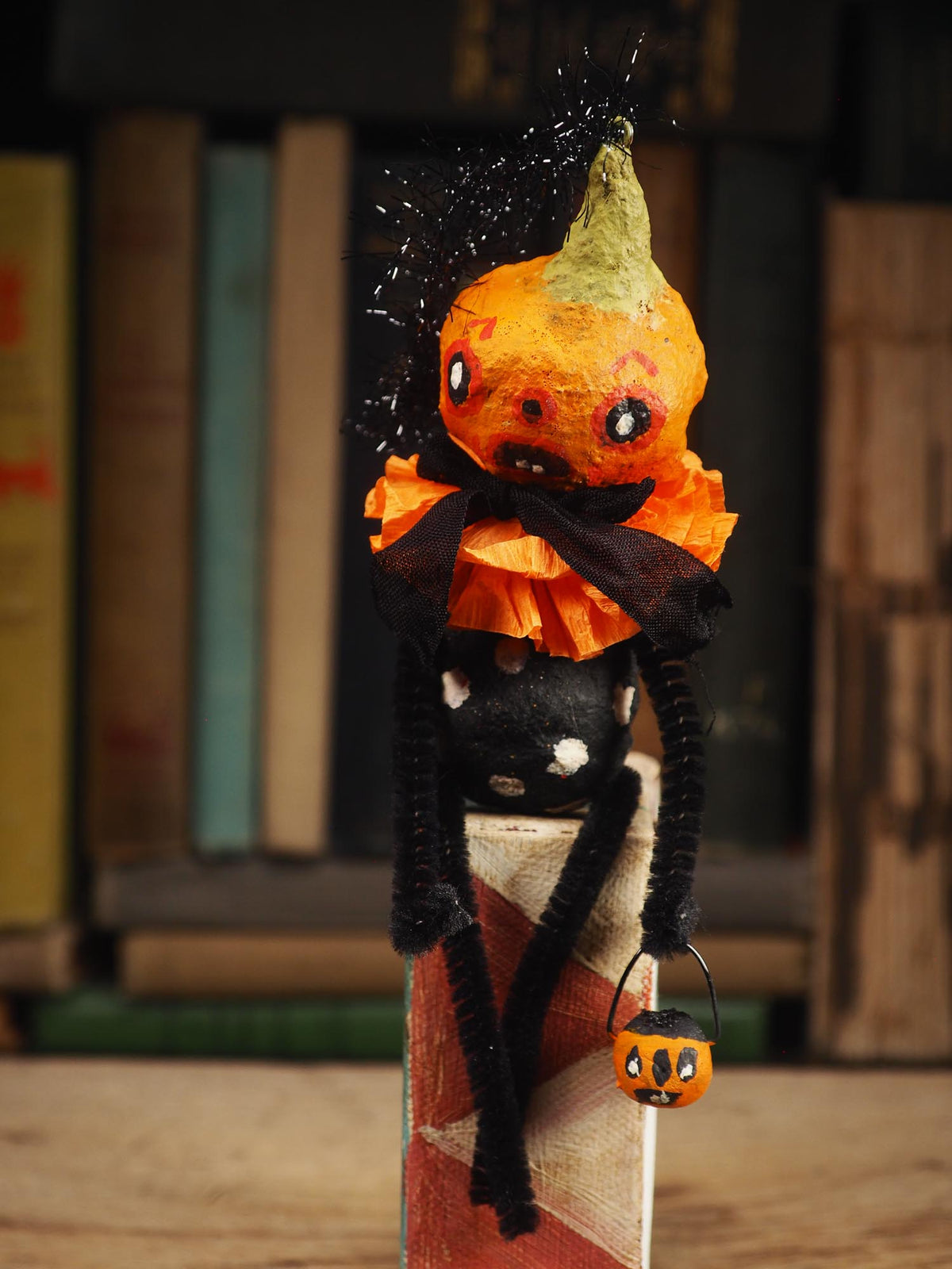 A handmade Halloween decoration shaped like a small orange jack-o'-lantern with a black polka-dotted body, orange ruffled collar, black pipe cleaners for arms, and a tiny orange pumpkin bucket, standing in front of a blurred bookshelf background. Original spun cotton hand made doll by Idania Salcido (Danita Art)