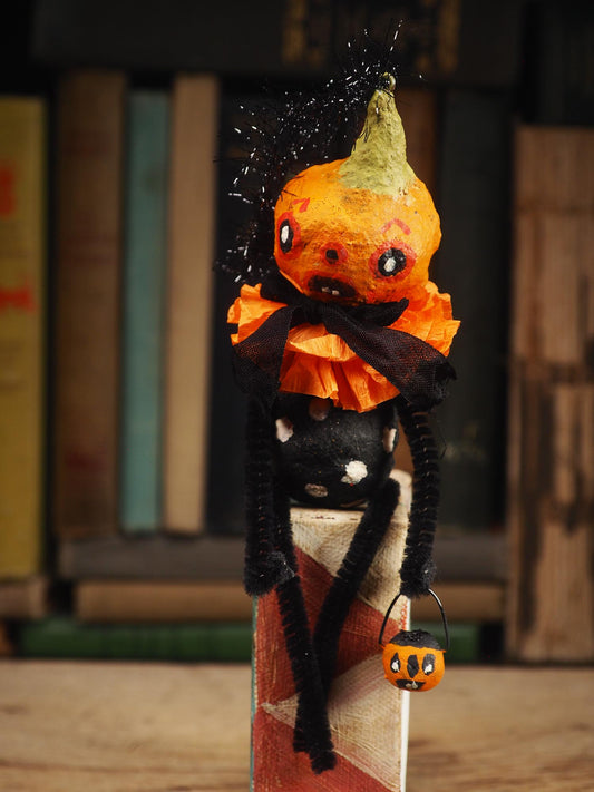A handmade Halloween decoration shaped like a small orange jack-o'-lantern with a black polka-dotted body, orange ruffled collar, black pipe cleaners for arms, and a tiny orange pumpkin bucket, standing in front of a blurred bookshelf background. Original spun cotton hand made doll by Idania Salcido (Danita Art)
