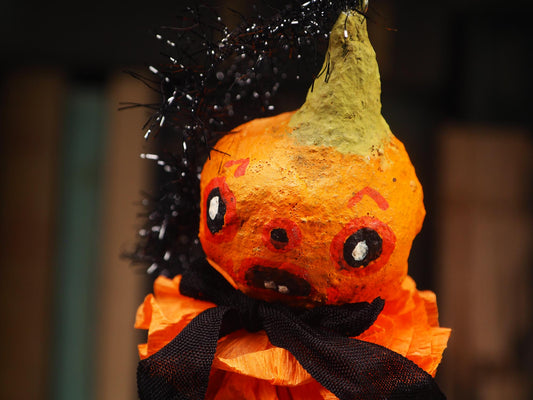 A close-up of a Halloween decoration shaped like a small orange pumpkin with a distressed texture, featuring a green stem, black tinsel hair, a black ribbon bow, and an orange ruffled skirt against a dark blurred background. Original spun cotton hand made Halloween doll by Idania Salcido (Danita Art)