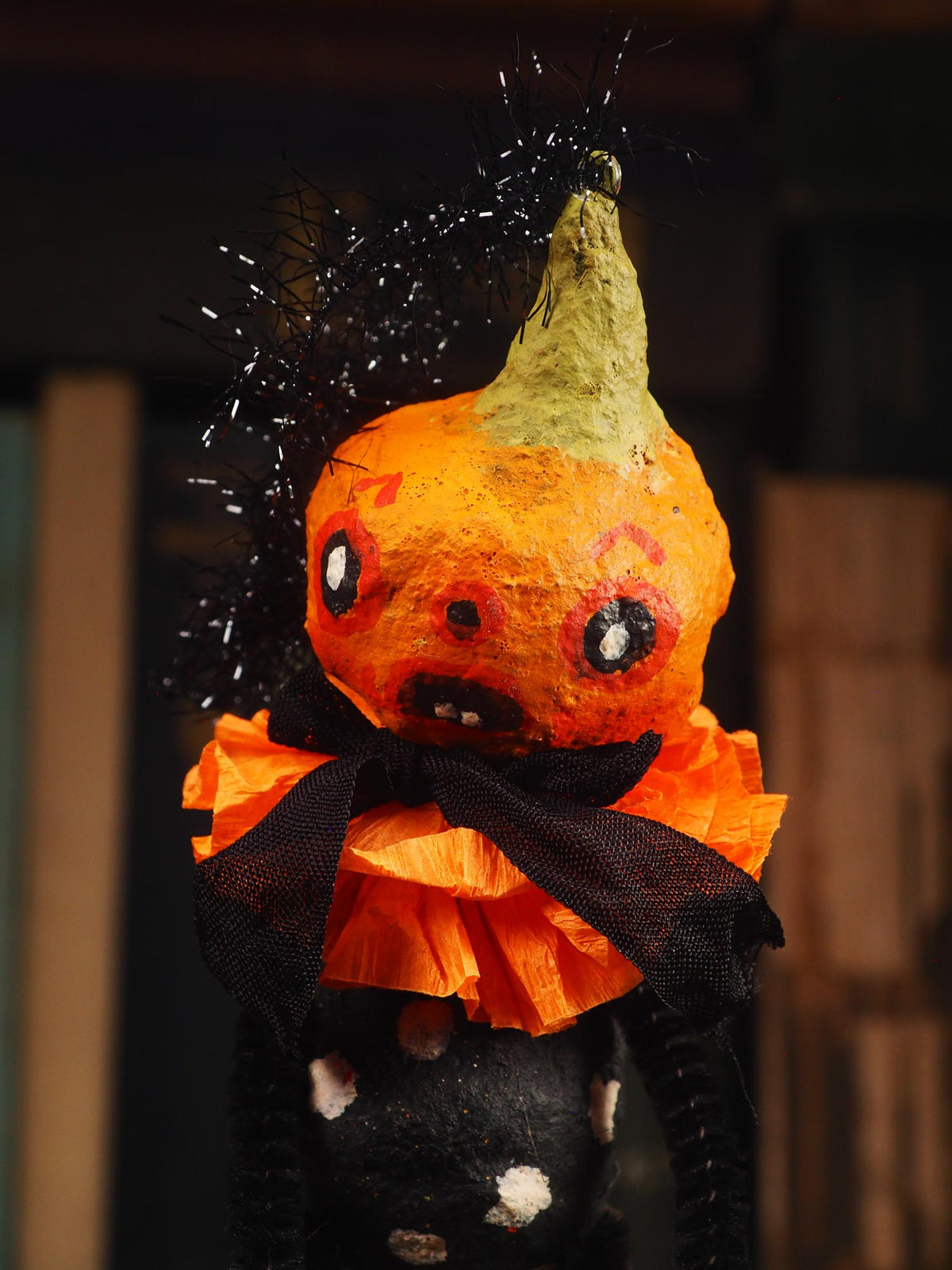 A Halloween decorative figure shaped like a distorted orange pumpkin with a green stem, black tinsel hair, a black bow, and an orange tissue-paper ruffled collar, shown against a dark blurred background. Original spun cotton hand made Halloween doll by Idania Salcido (Danita Art)