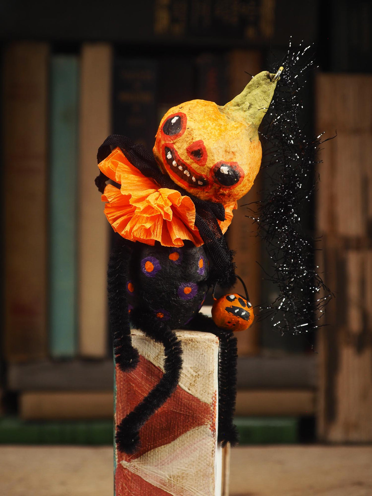 A Halloween decorative spun cotton figure shaped like a small orange pumpkin head with a black body, orange ruffled collar, black fuzzy limbs, and a green hat, posed on top of a book with a blurred bookshelf background. Hand Made By Idania Salcido (Danita Art)