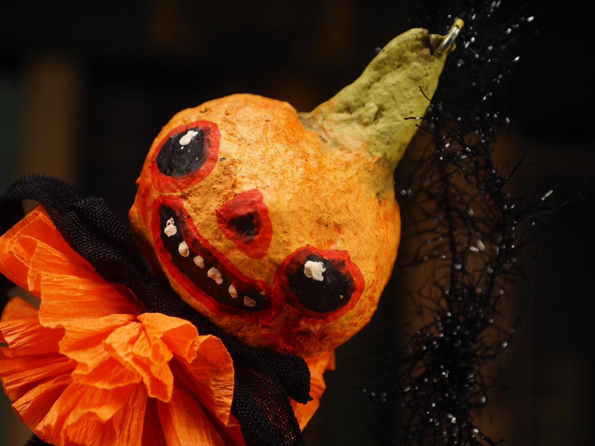A close-up of a spun cotton decorative orange gourd Halloween ornament with a painted scary face, black eye patches, a green stem, an orange tissue-paper ruffle, and black tinsel against a dark background. Handmade by Idania Salcido (Danita Art)