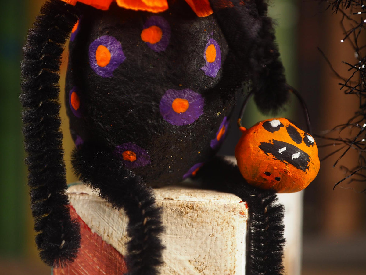 A close-up of a spun cotton decorative orange gourd Halloween ornament with a painted scary face, black eye patches, a green stem, an orange tissue-paper ruffle, and black tinsel against a dark background. Handmade by Idania Salcido (Danita Art)
