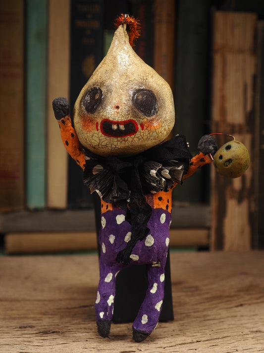 A handmade folk art doll shaped like a cracked beige gourd head with black eyes and a red mouth, wearing an orange and black polka-dot top and purple pants with white dots, standing on a wooden surface with a blurred bookshelf background. An original hand made Halloween art doll by Idania Salcido AKA Danita Art.