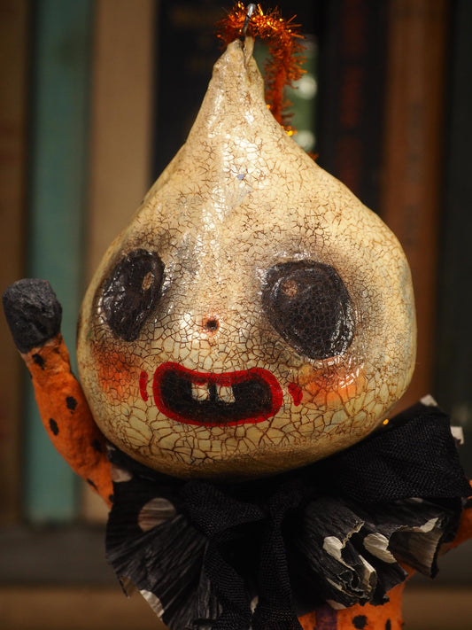 A handmade folk art doll shaped like a cracked beige gourd head with black eyes and a red mouth, wearing an orange and black polka-dot top and purple pants with white dots, standing on a wooden surface with a blurred bookshelf background. An original hand made Halloween art doll by Idania Salcido AKA Danita Art.
