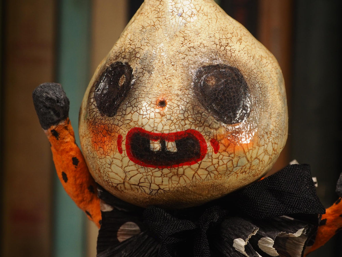 A handmade folk art doll shaped like a cracked beige gourd head with black eyes and a red mouth, wearing an orange and black polka-dot top and purple pants with white dots, standing on a wooden surface with a blurred bookshelf background. An original hand made Halloween art doll by Idania Salcido AKA Danita Art.
