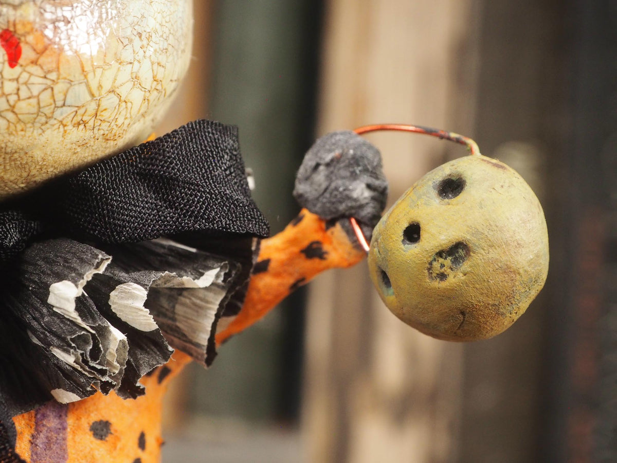 A handmade folk art doll shaped like a cracked beige gourd head with black eyes and a red mouth, wearing an orange and black polka-dot top and purple pants with white dots, standing on a wooden surface with a blurred bookshelf background. An original hand made Halloween art doll by Idania Salcido AKA Danita Art.